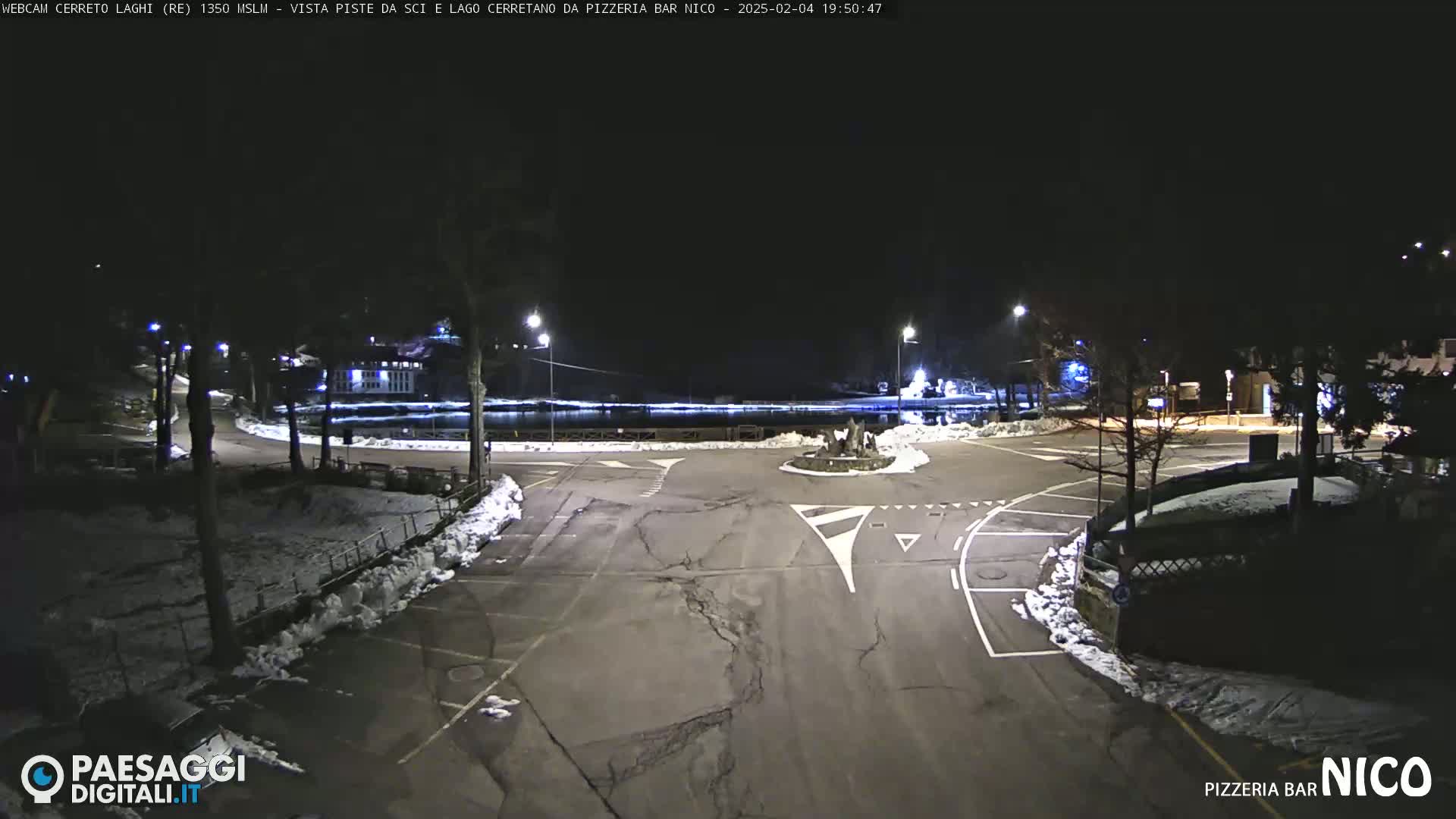 A nighttime view of a snow-dusted parking lot and roundabout near a body of water, illuminated by streetlights.