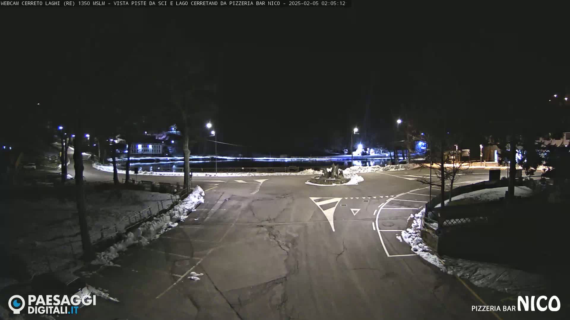 A nighttime view of a snow-dusted parking lot and roadway intersection, illuminated by streetlights.