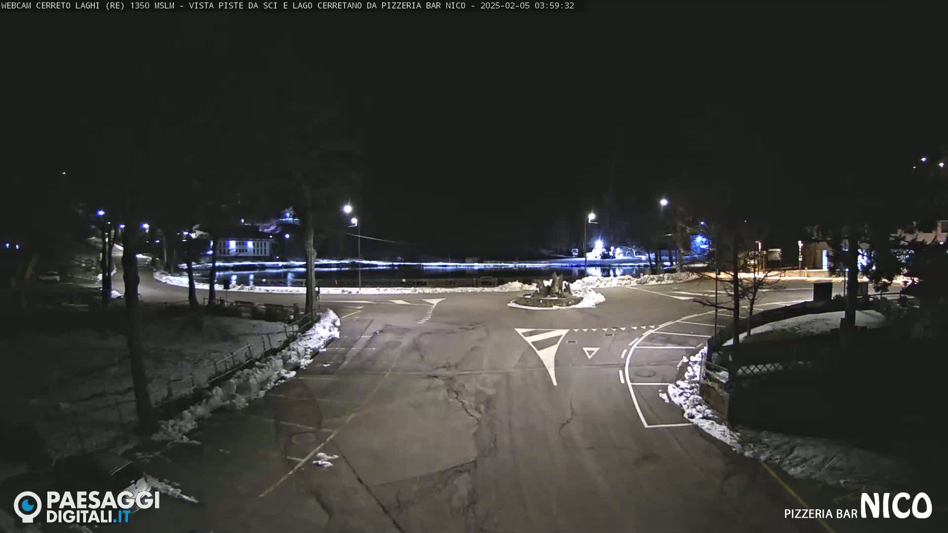 A nighttime view of a snow-dusted roundabout and parking lot near a lake, illuminated by streetlights.