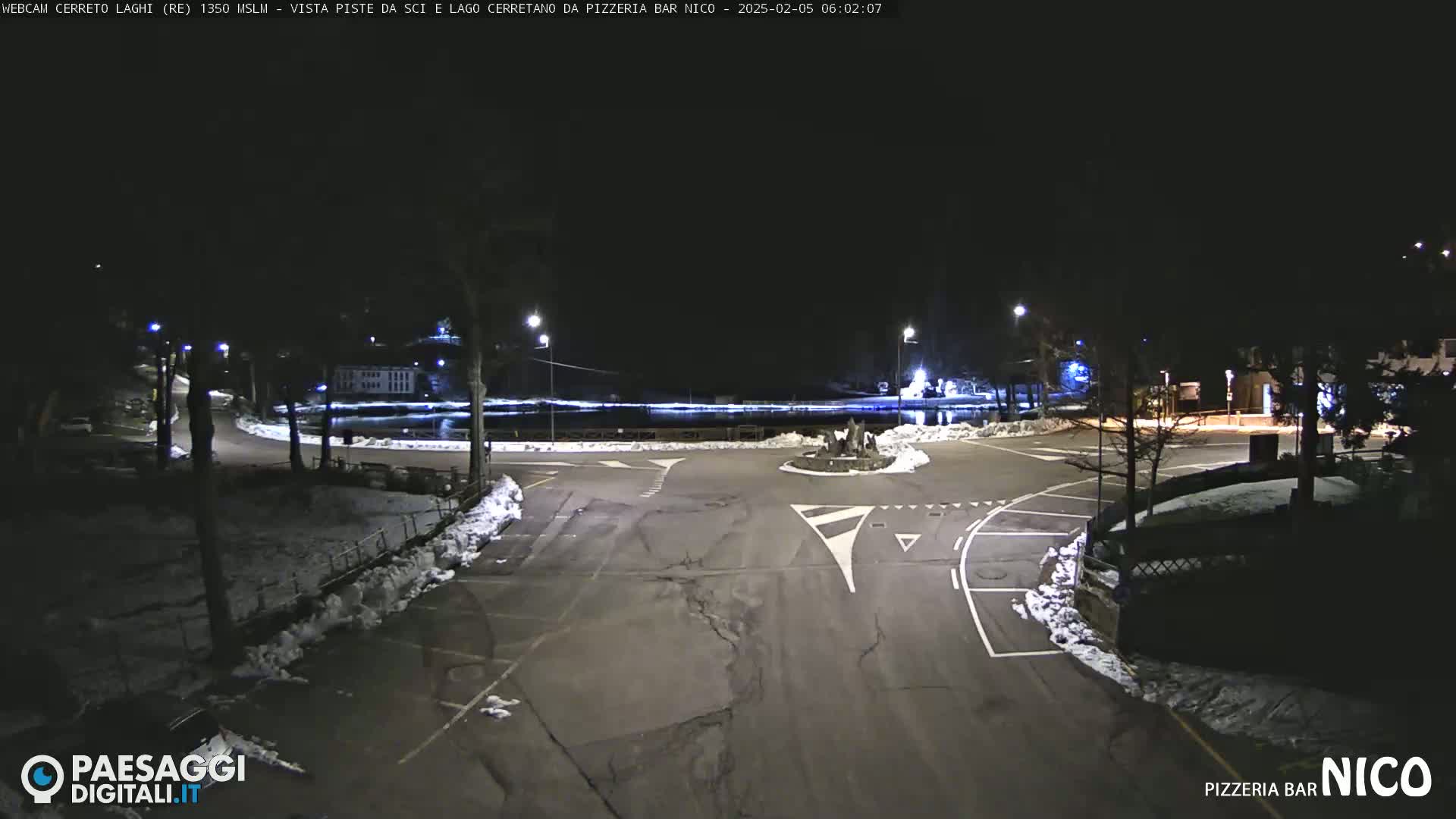 A nighttime view of a snow-dusted parking lot and roadway intersection, illuminated by streetlights, with a body of water visible in the background.