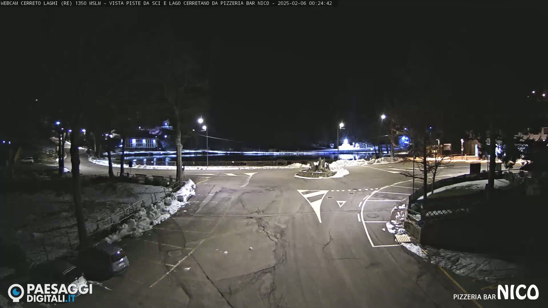 A nighttime view of a snow-dusted parking lot and roadway intersection near a body of water, illuminated by streetlights.