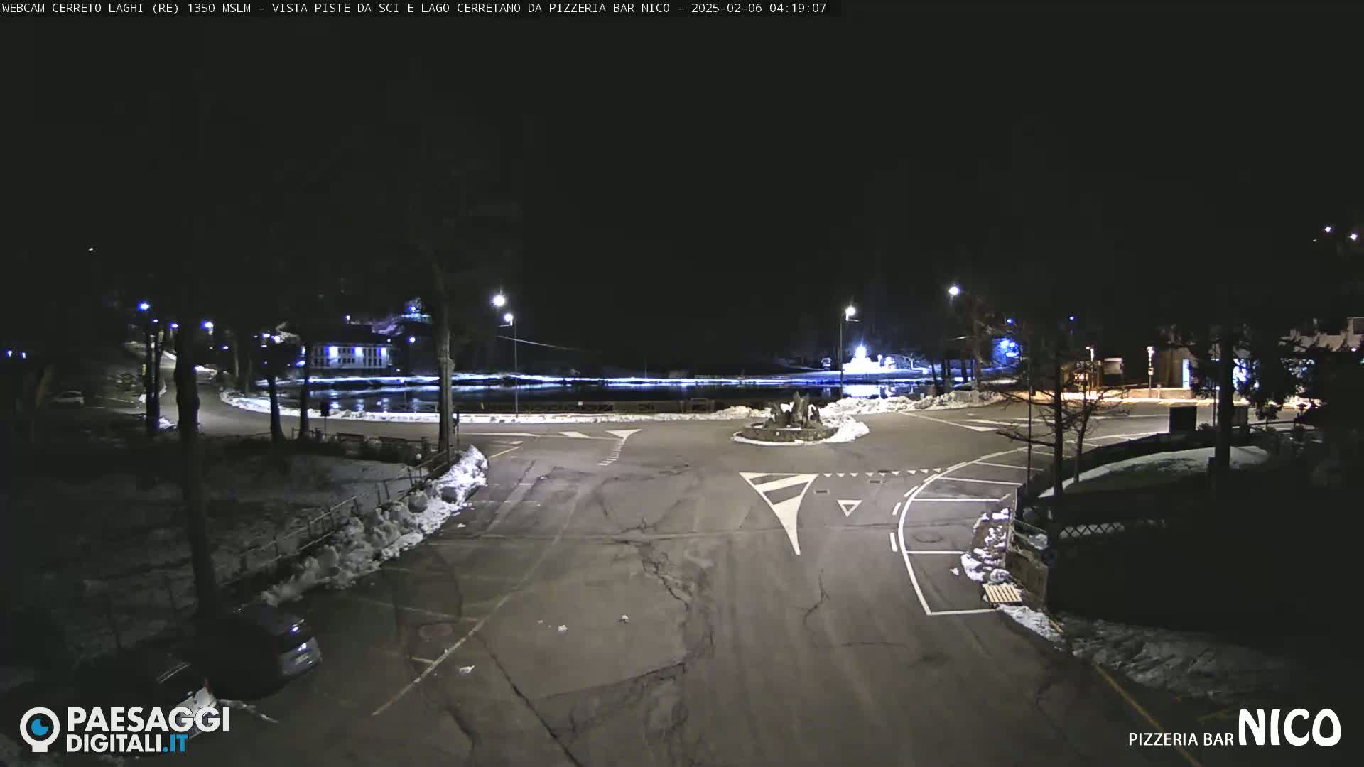 A nighttime, snow-dusted parking lot and roadway with some nearby buildings and a body of water, illuminated by streetlights.