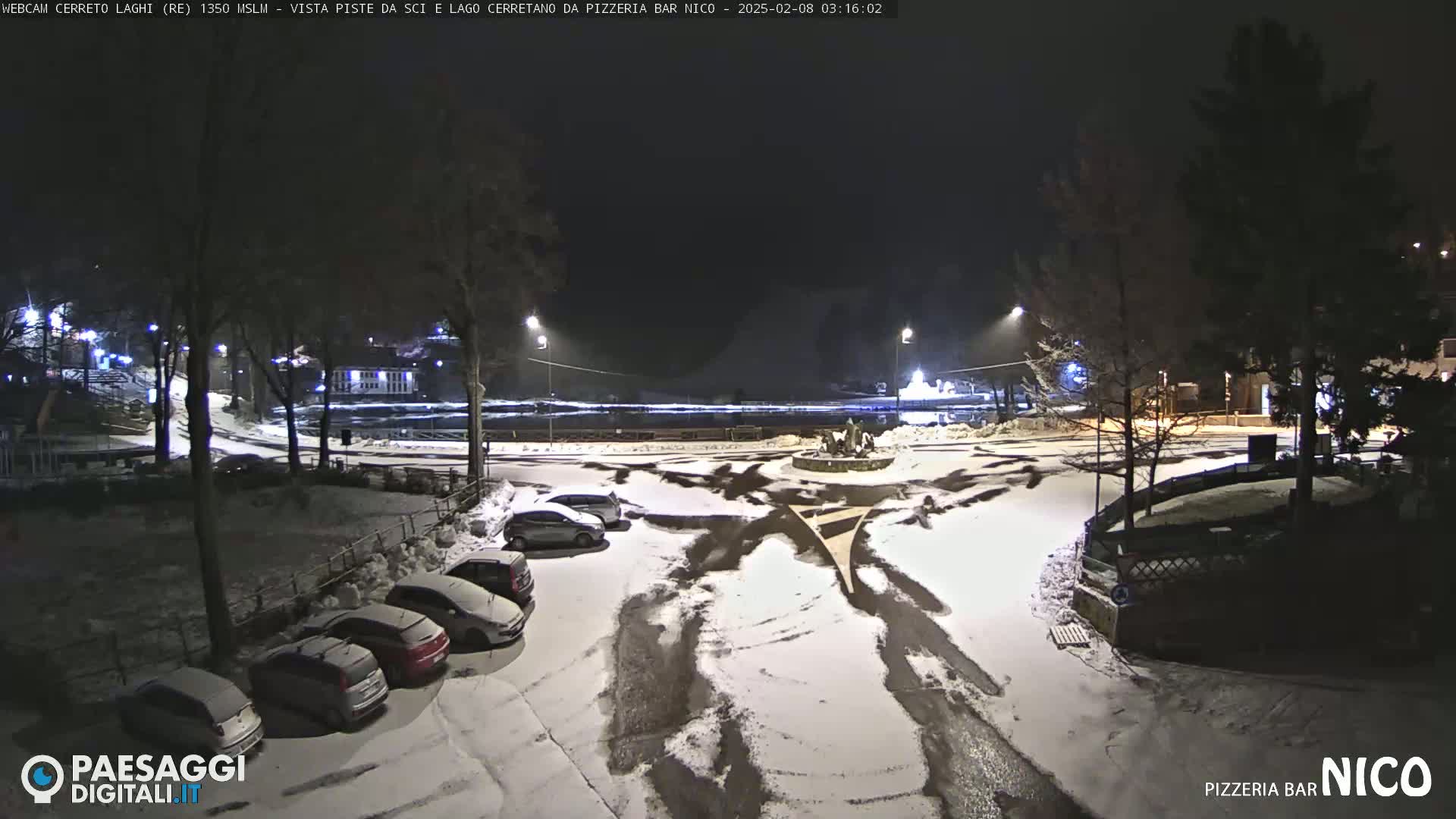 A snow-covered parking lot at night, with several cars parked and streetlights illuminating the scene.