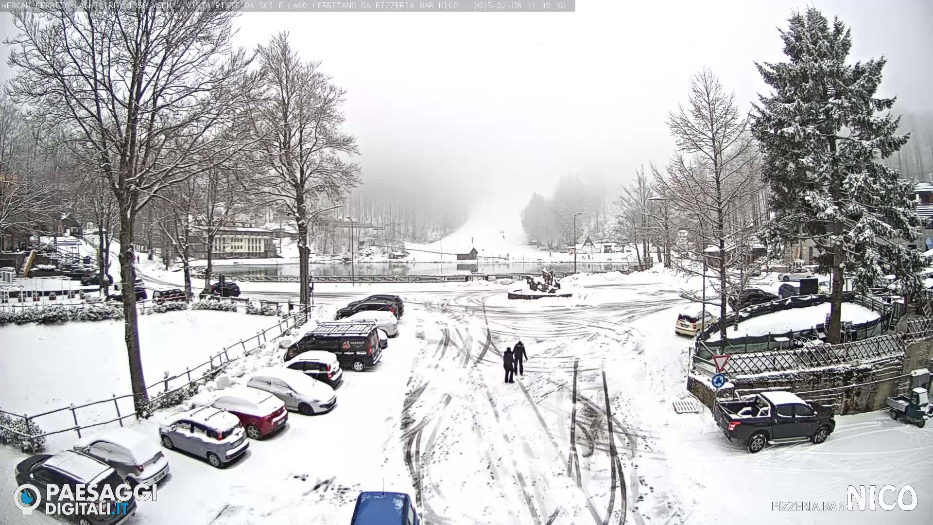 A snow-covered parking lot and roadway are surrounded by snow-laden trees and buildings, with two people walking in the distance during a snowfall.