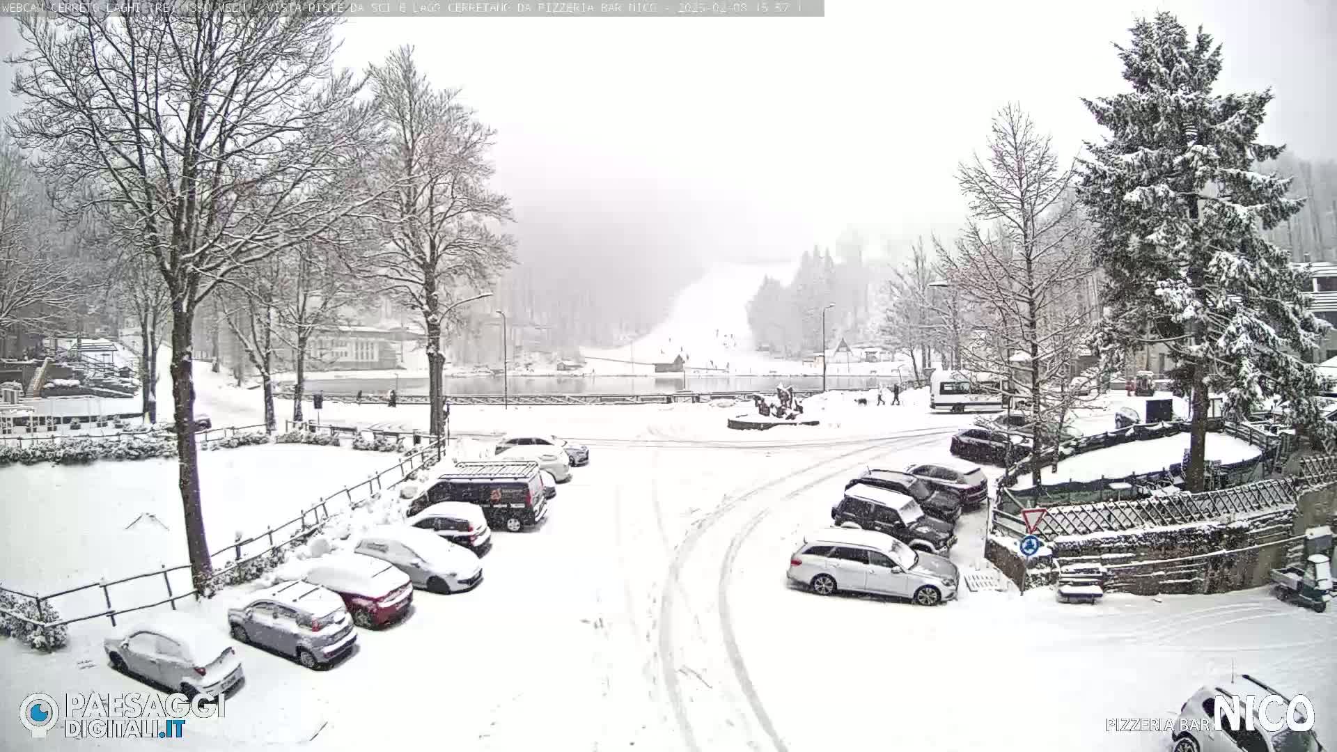 A snow-covered parking lot and town square with cars parked, and bare trees covered in snow under a heavy snowfall.