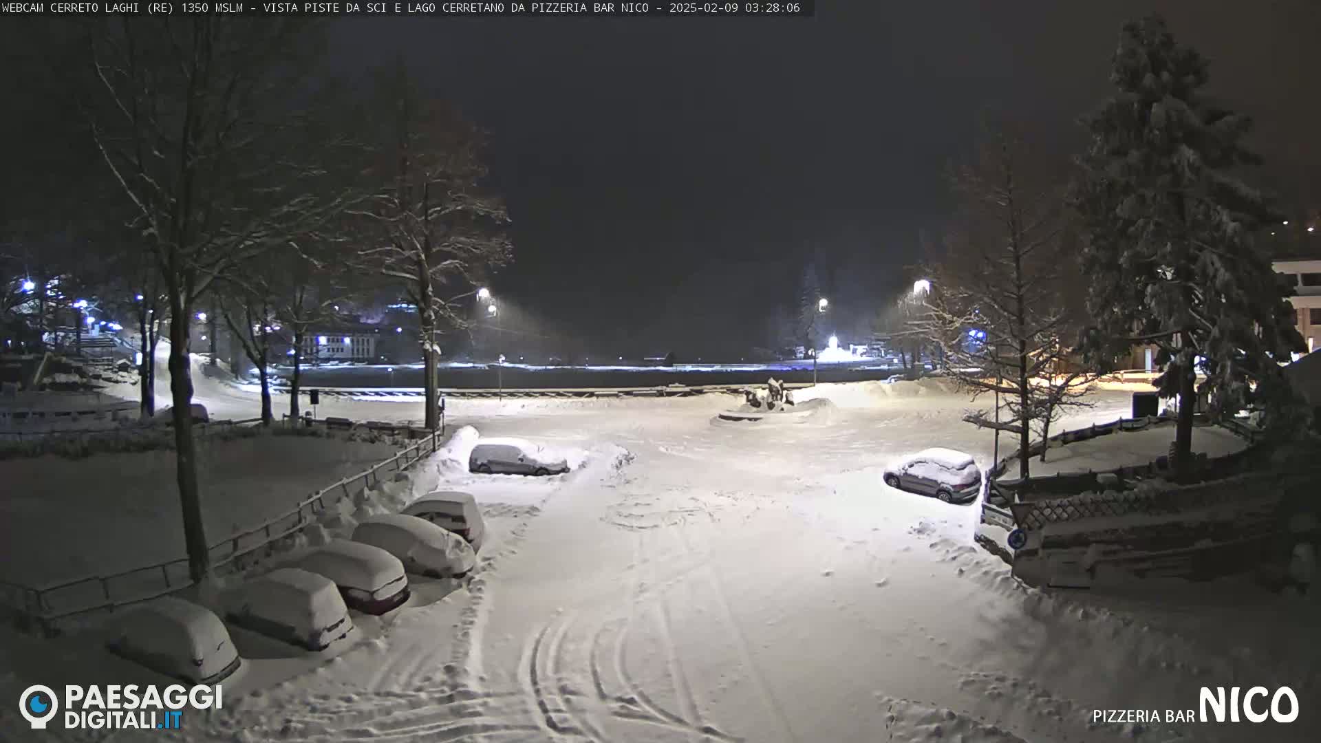 A snow-covered parking lot at night, with several cars parked and streetlights illuminating the scene.
