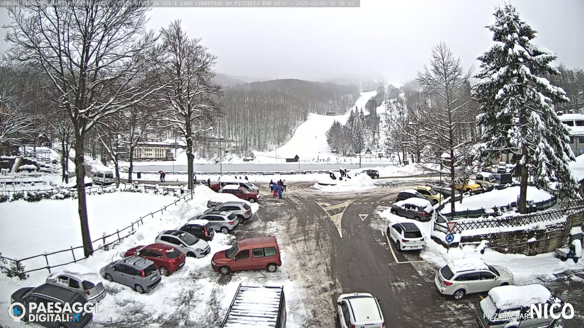 A snow-covered parking lot and road are surrounded by snow-covered trees and buildings, with a ski slope visible in the background under a cloudy sky.