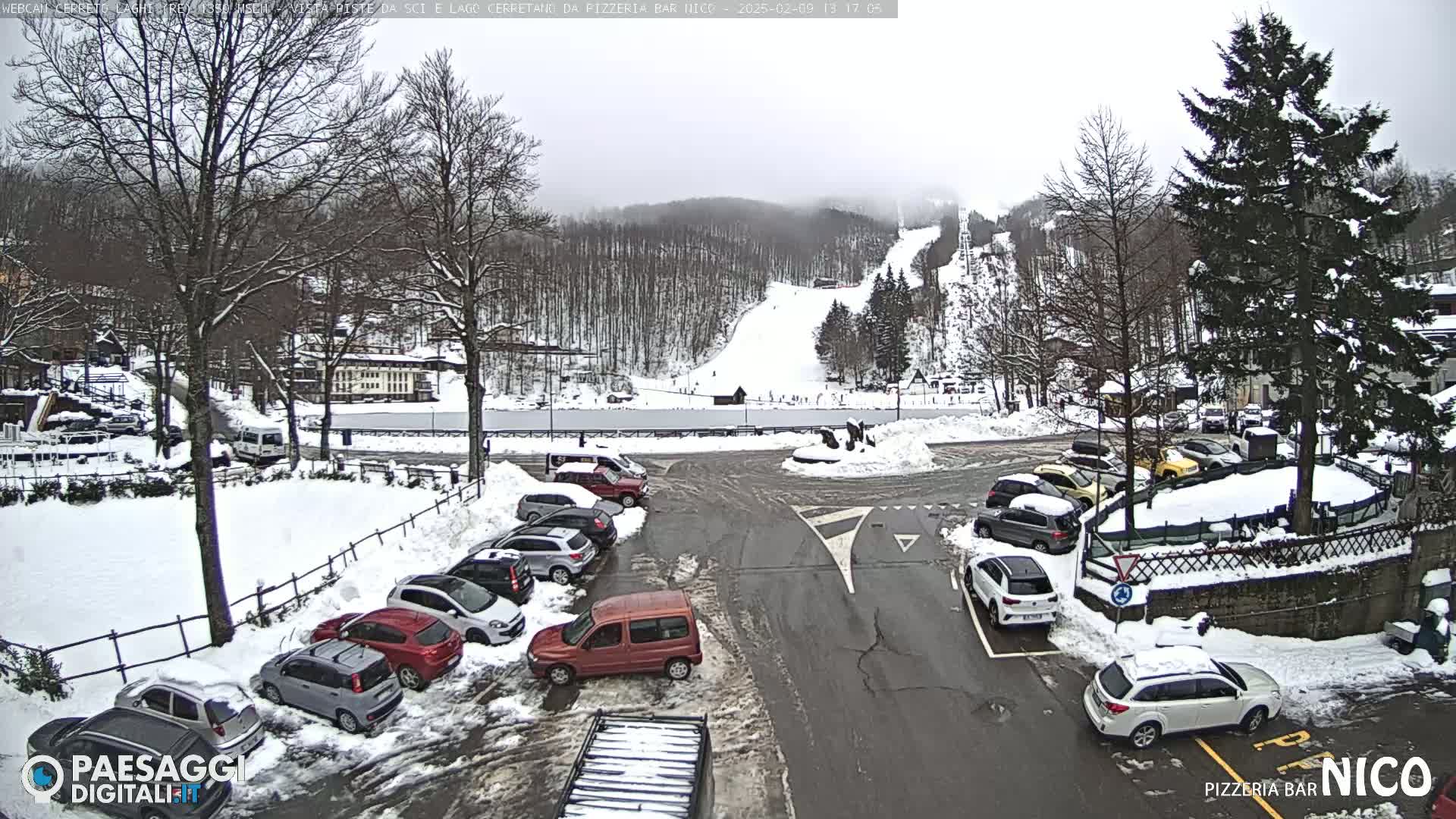 A snow-covered parking lot with cars and a view of a snowy ski slope in the background under an overcast sky.
