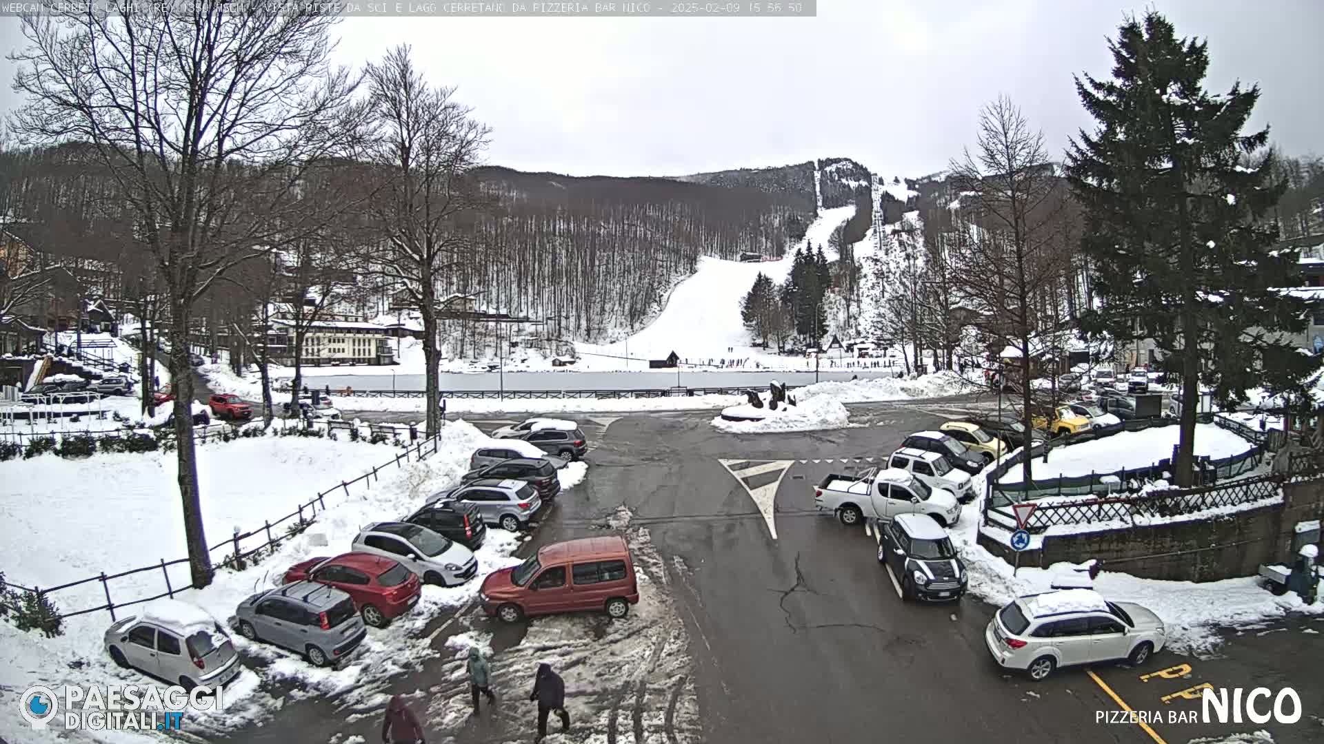A snow-covered parking lot with cars and a ski slope in the background on an overcast day.