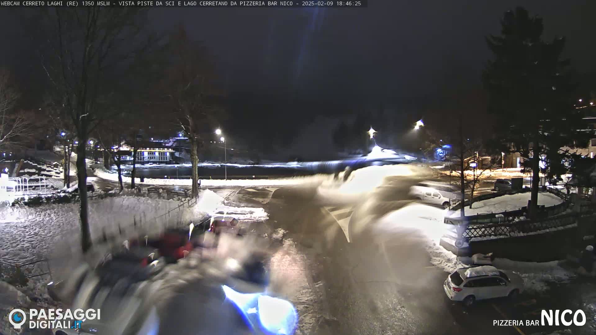 A nighttime snow scene shows a snowplow clearing a roadway near parked cars and buildings under streetlights.