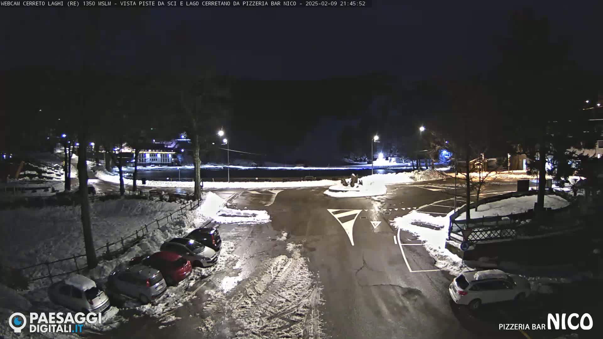 A nighttime, snow-covered parking lot with several cars parked, illuminated by streetlights, near a body of water.