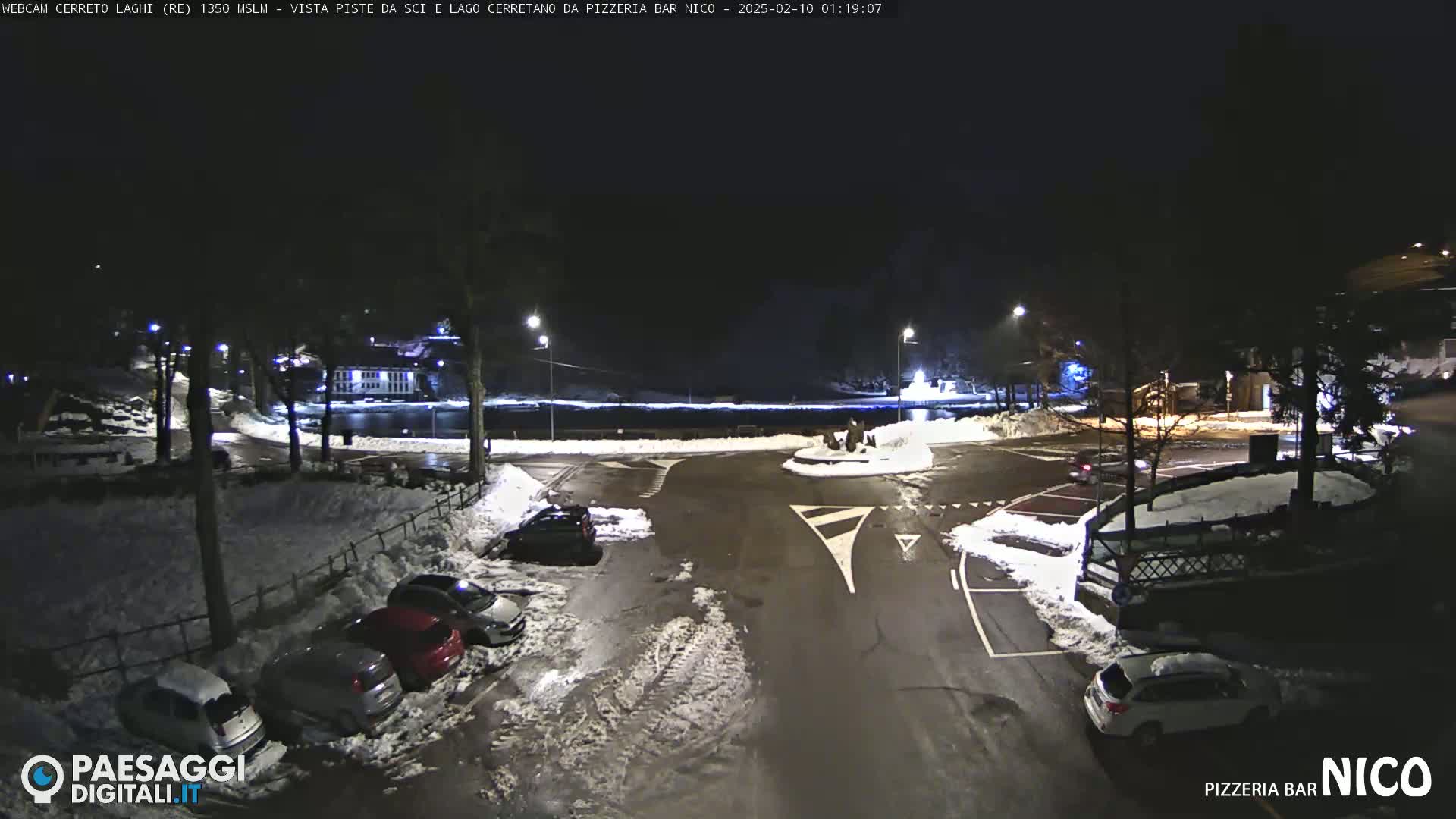 A nighttime scene of a snow-covered parking lot and roadway surrounding a roundabout, illuminated by streetlights.