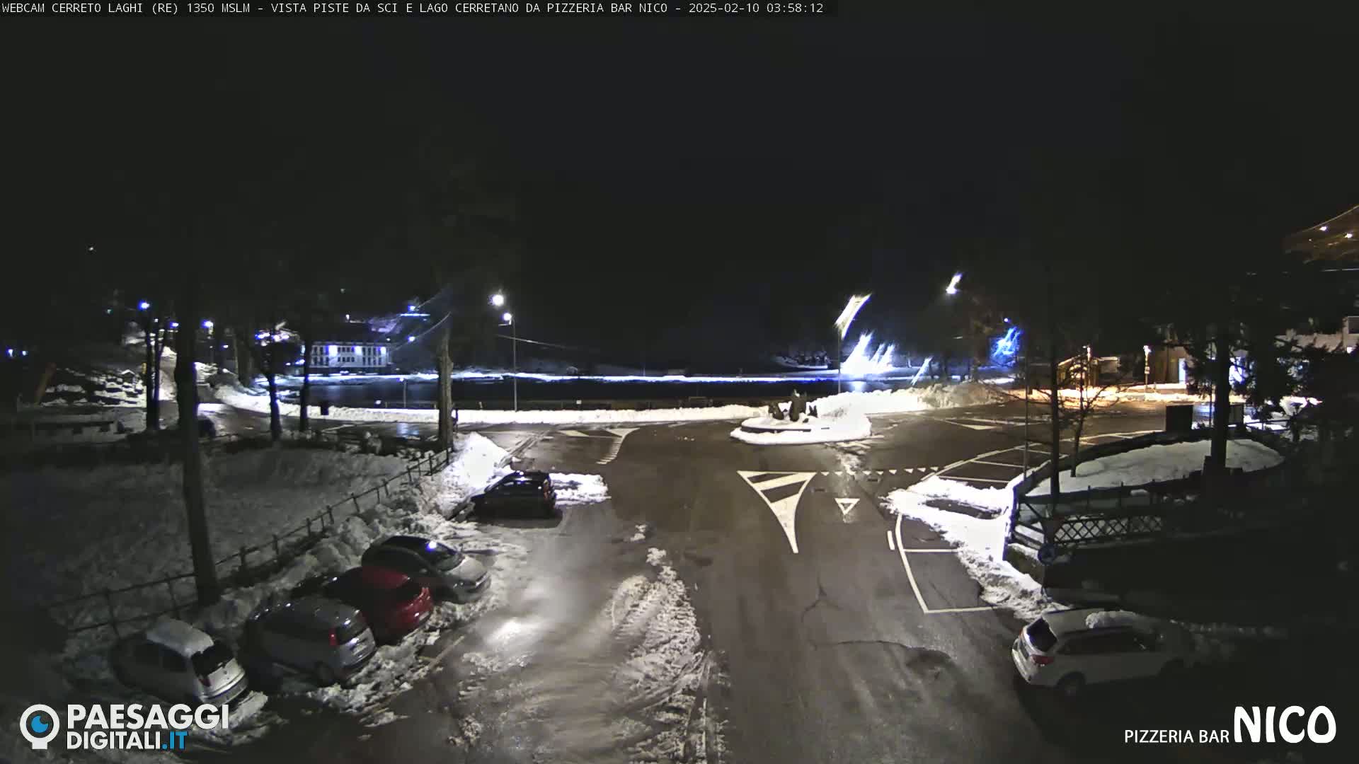 A nighttime, snow-covered parking lot with several cars and a building in the background.
