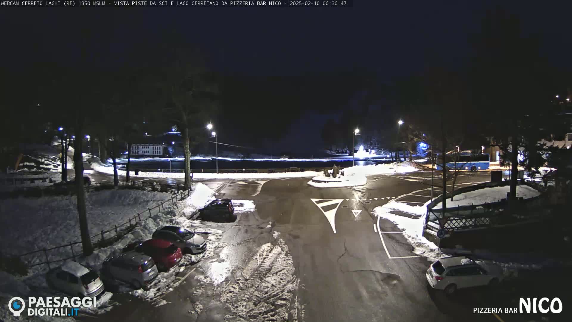 A snow-covered parking lot at night, illuminated by streetlights, with several cars parked and a body of water visible in the background.