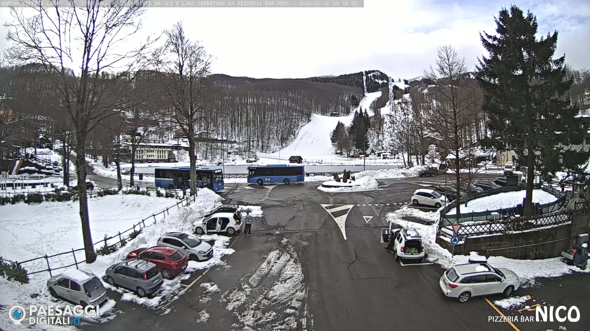 A snow-covered parking lot with several cars and a bus, situated near a ski slope and surrounded by trees under a cloudy sky.