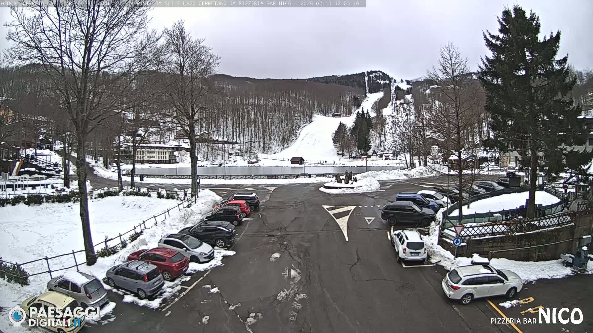 A snow-covered parking lot with cars and a background view of a ski slope on a cloudy day.