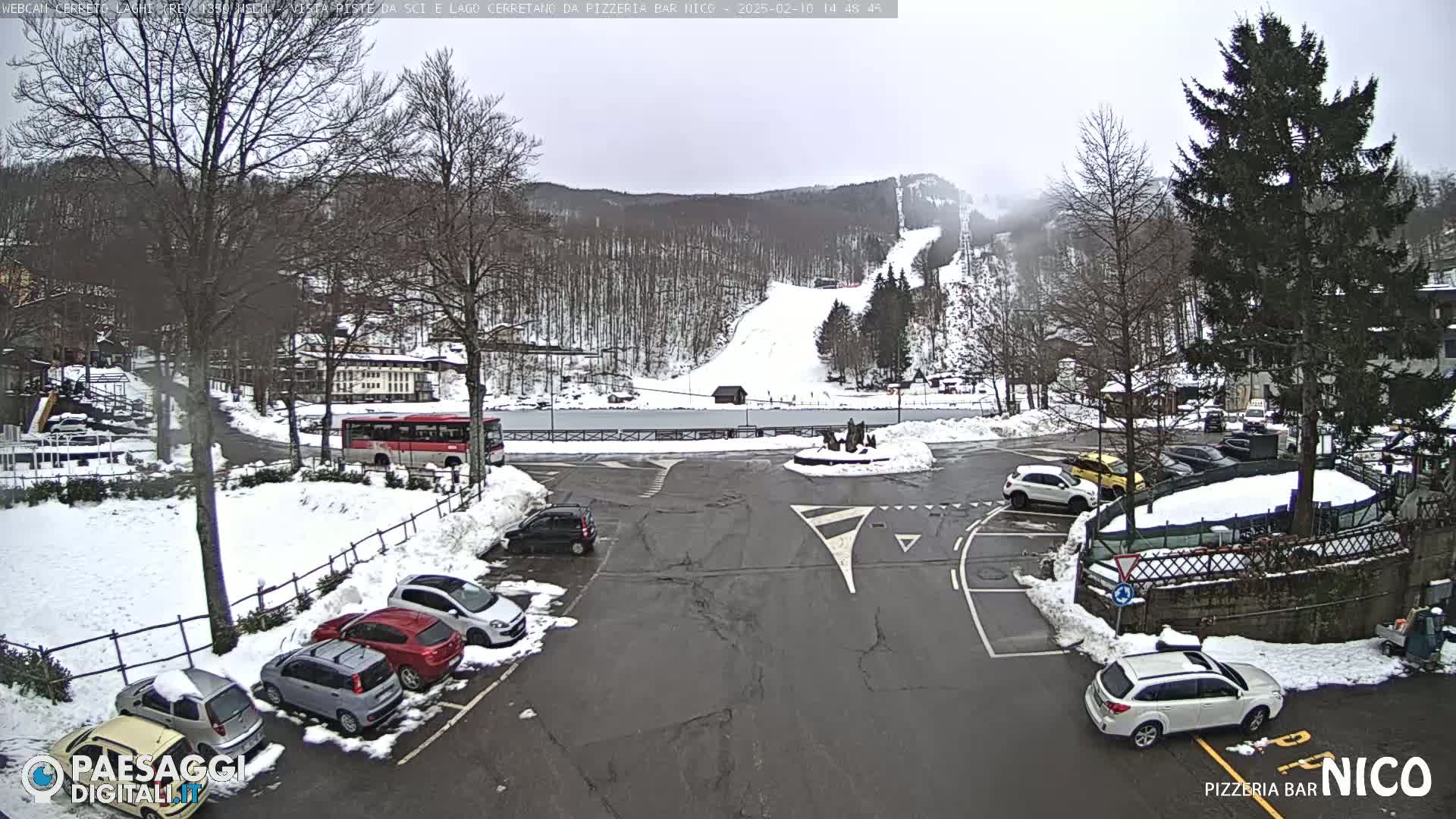 A snow-covered parking lot with several cars and a bus, situated near a ski slope and a body of water, under an overcast sky.
