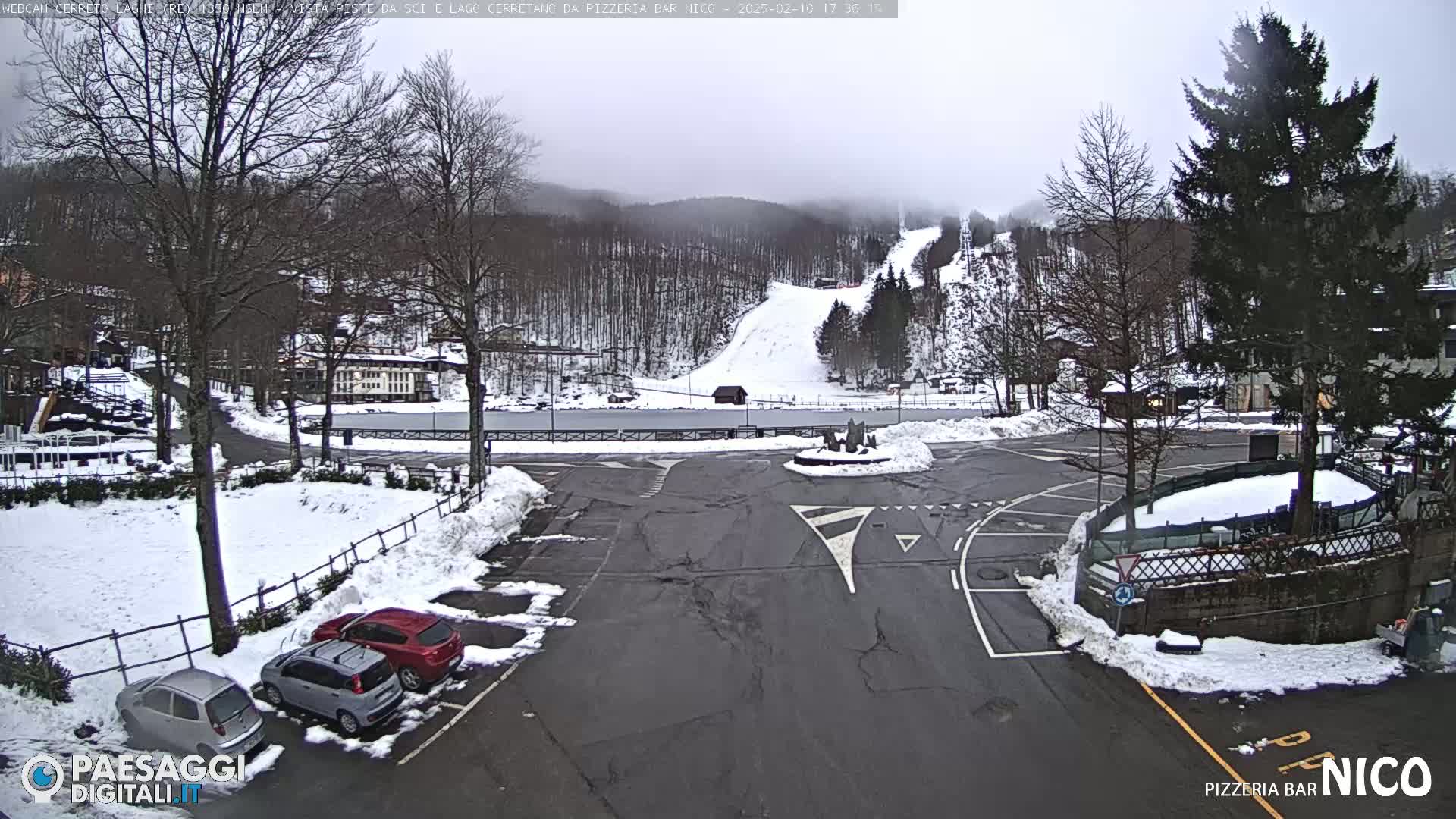 A snow-covered parking lot with several cars and a view of a ski slope in the background under an overcast sky.