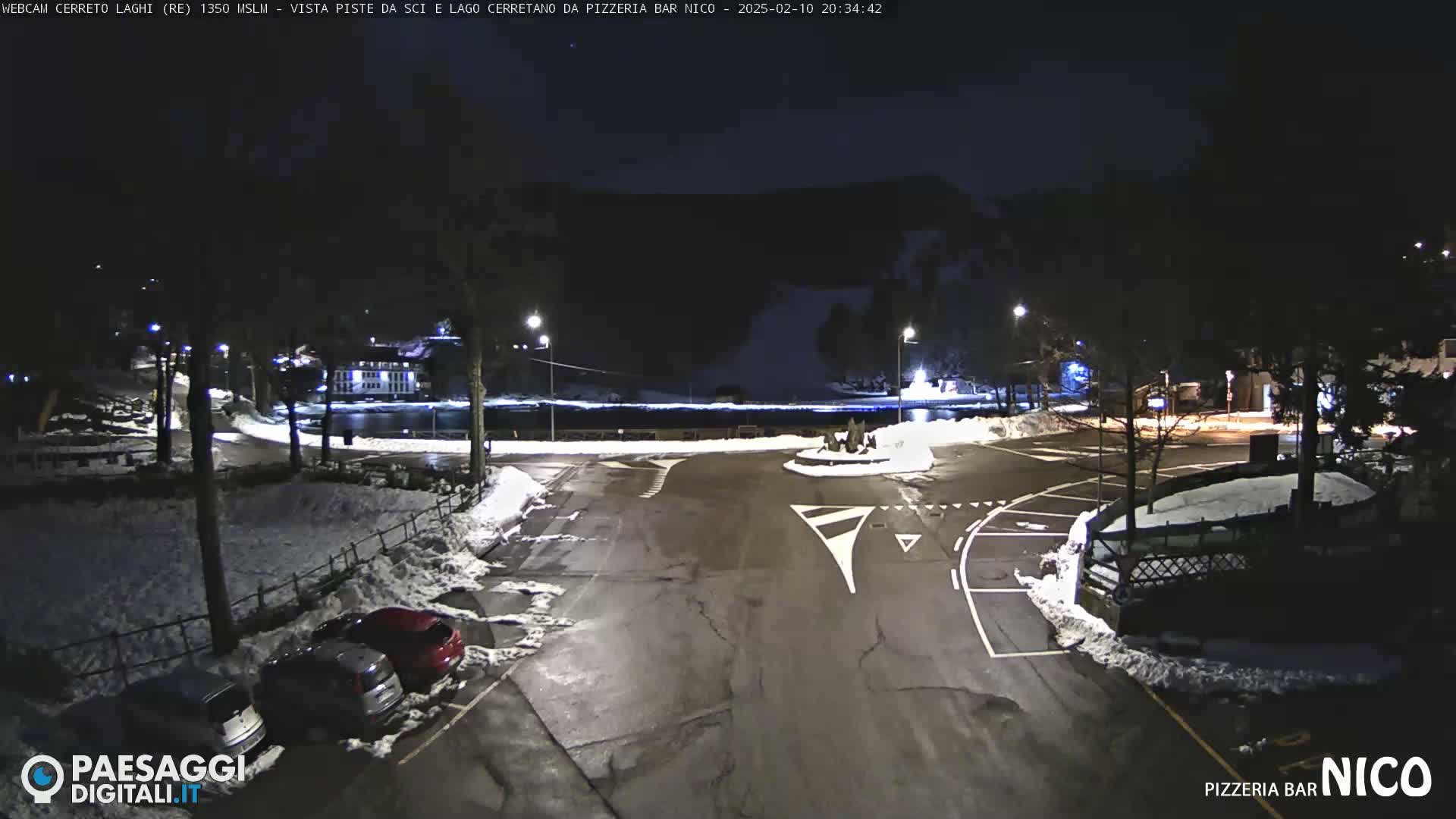 A nighttime view of a snow-covered parking lot and roadway near a body of water, with some cars parked and streetlights illuminating the scene.