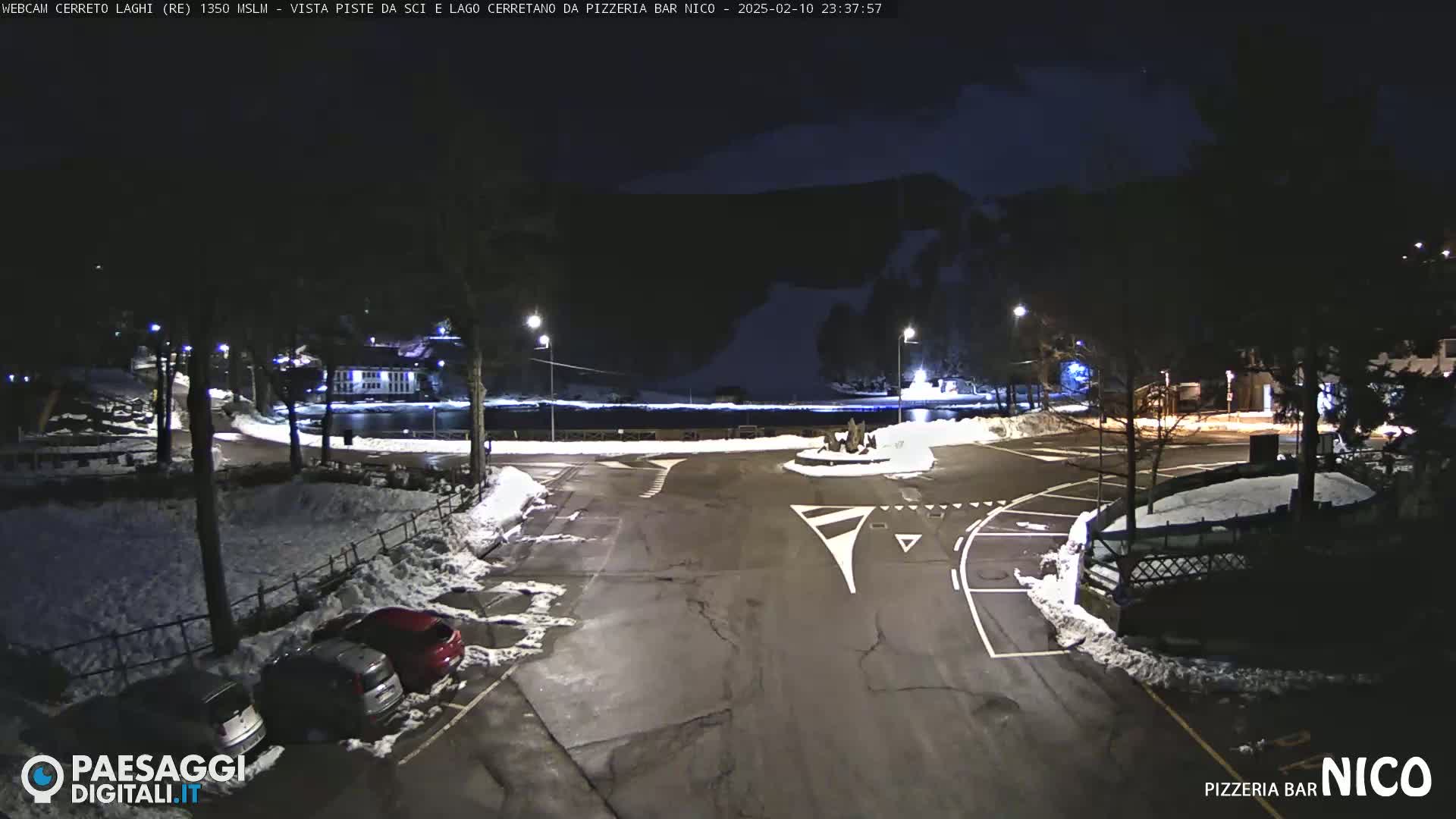 A nighttime parking lot with some cars, partially covered in snow, near a circular road and a body of water under a dark, clear sky.