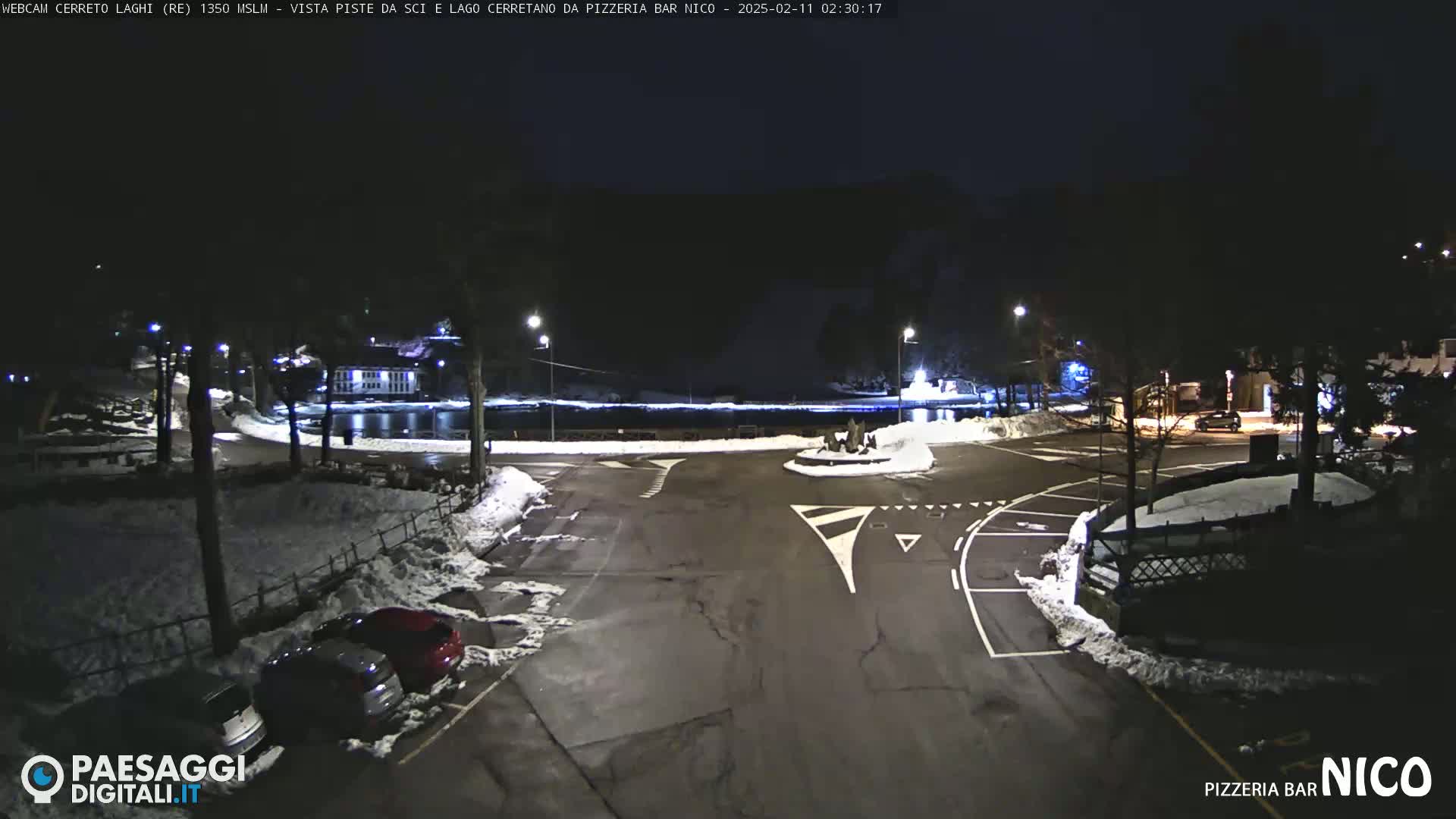 A nighttime view of a snow-covered parking lot and roadway surrounding a small body of water, illuminated by streetlights.