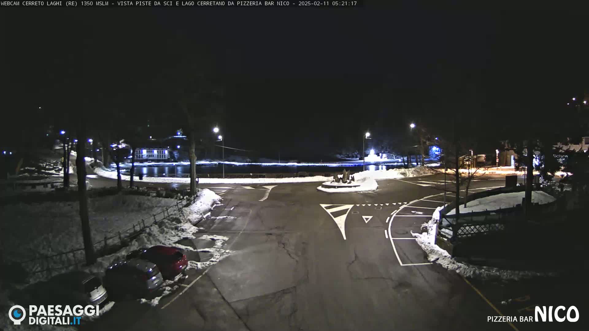 A nighttime view of a snow-covered parking lot and roadway curving around a small body of water, illuminated by streetlights.