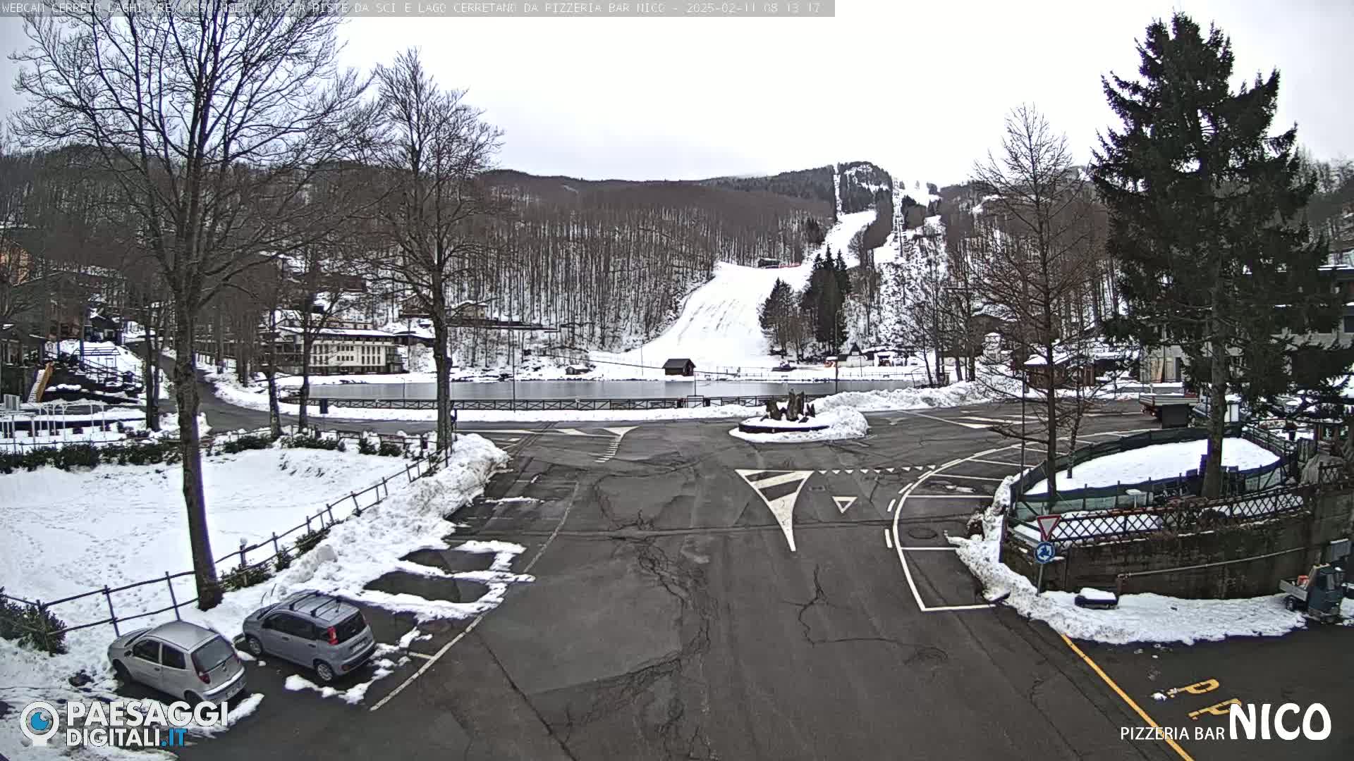 A snow-covered parking lot and roadway are situated in front of a snow-dusted ski slope and wooded hillside under an overcast sky.