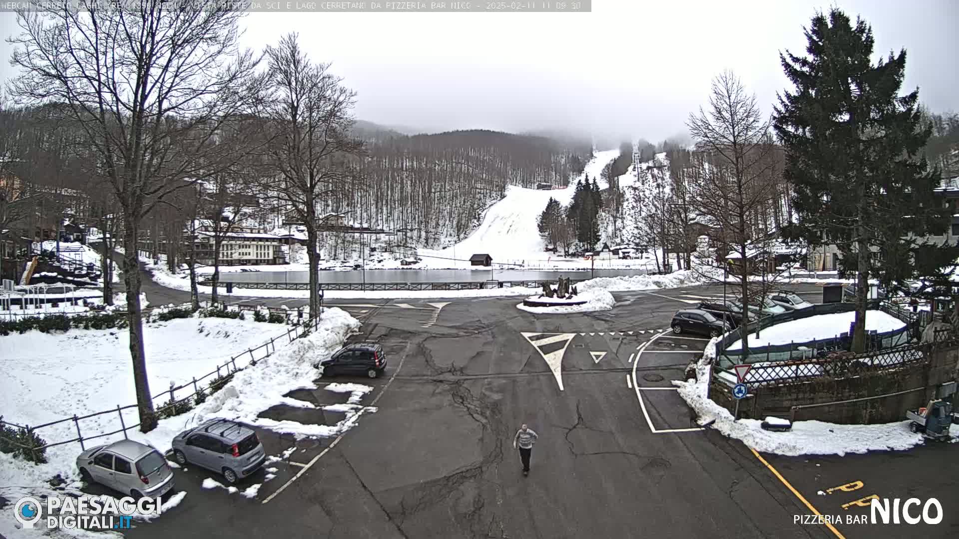 A snowy parking lot and roadway are surrounded by snow-covered trees and buildings, with a ski slope visible in the background under an overcast sky.