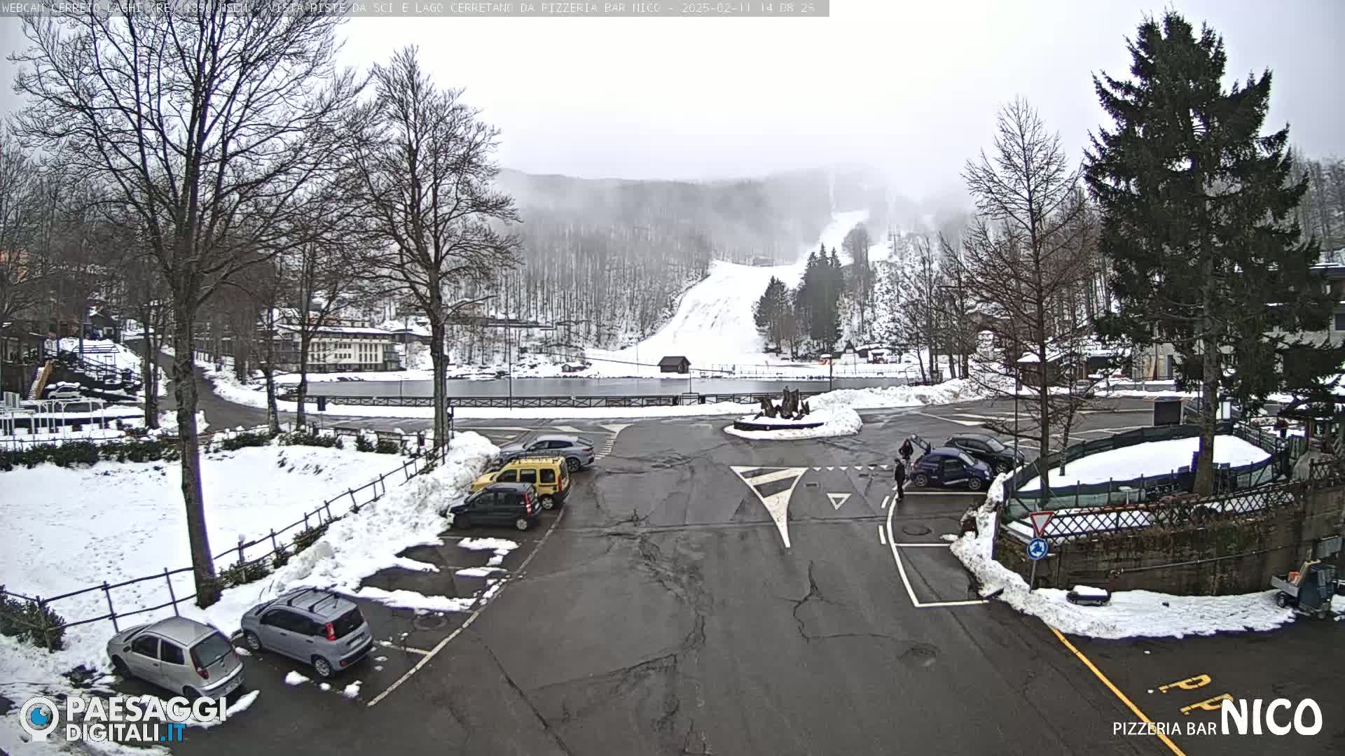 A snowy parking lot with several cars is situated near a frozen lake and a ski slope on a foggy day.