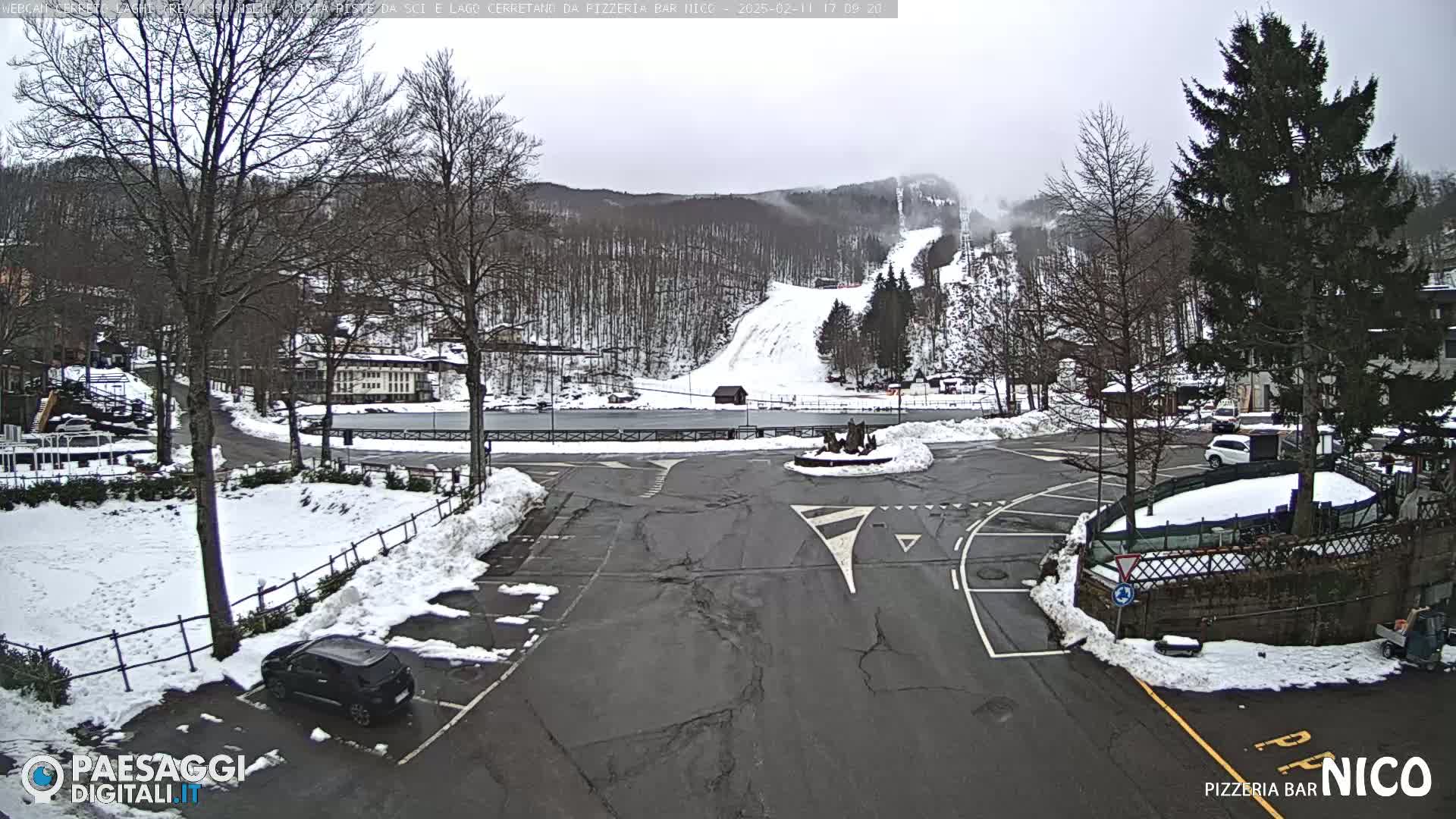 A snow-covered parking lot and roadway overlook a ski slope in a wooded area under an overcast sky.