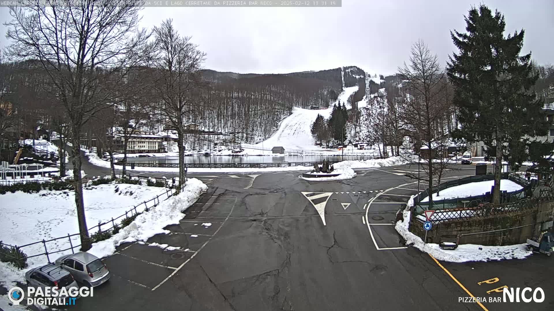 A snow-covered parking lot and roadway are situated in front of a partially snow-covered ski slope and wooded hillside under an overcast sky.