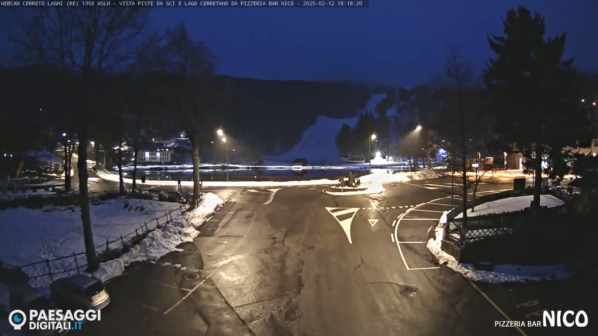 A dark, snow-dusted parking lot and roadway are illuminated at night, with a calm body of water and a hillside visible in the background under a twilight sky.