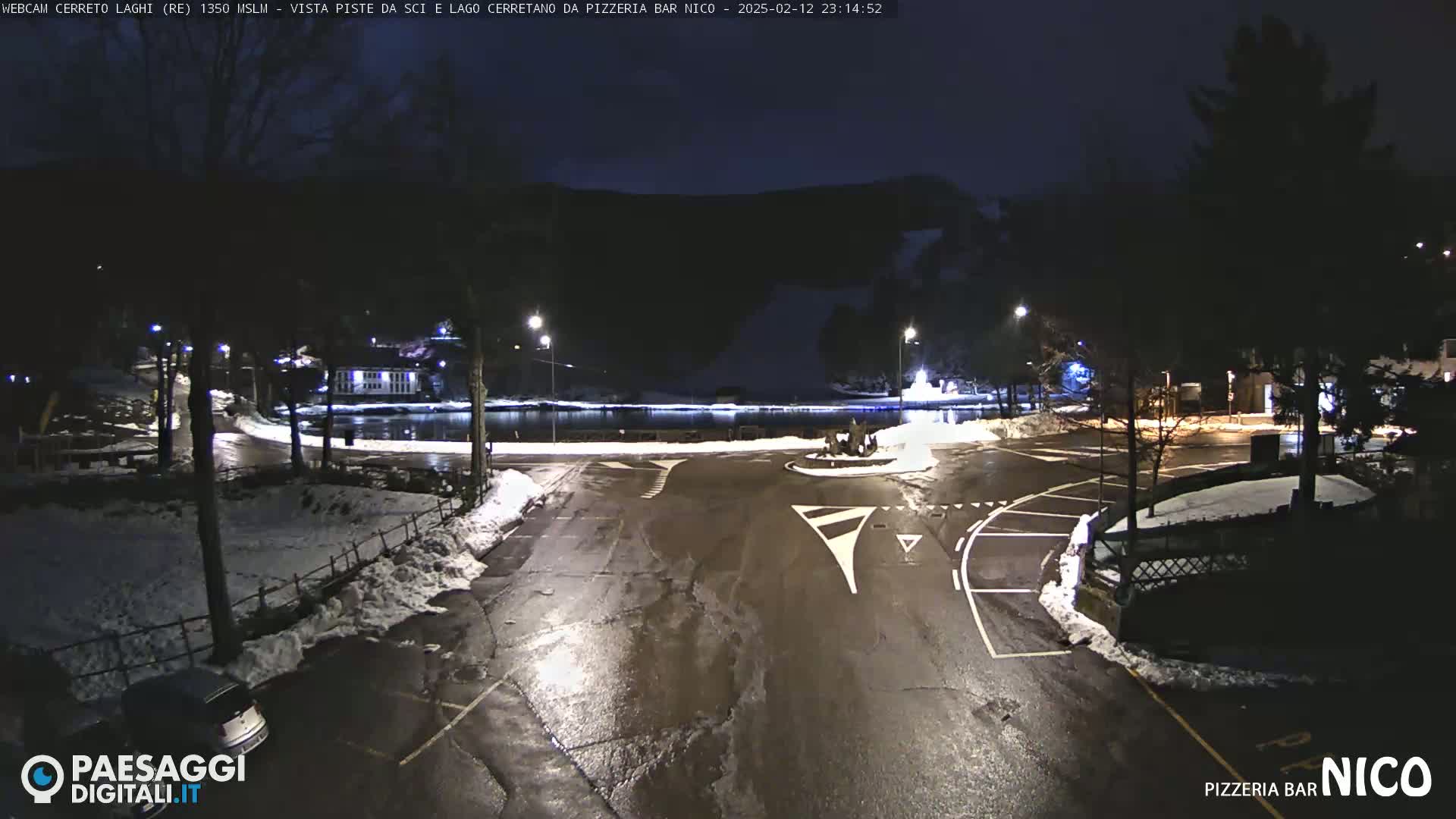 A nighttime scene of a snow-dusted parking lot and roadway near a calm body of water, under a dark sky.