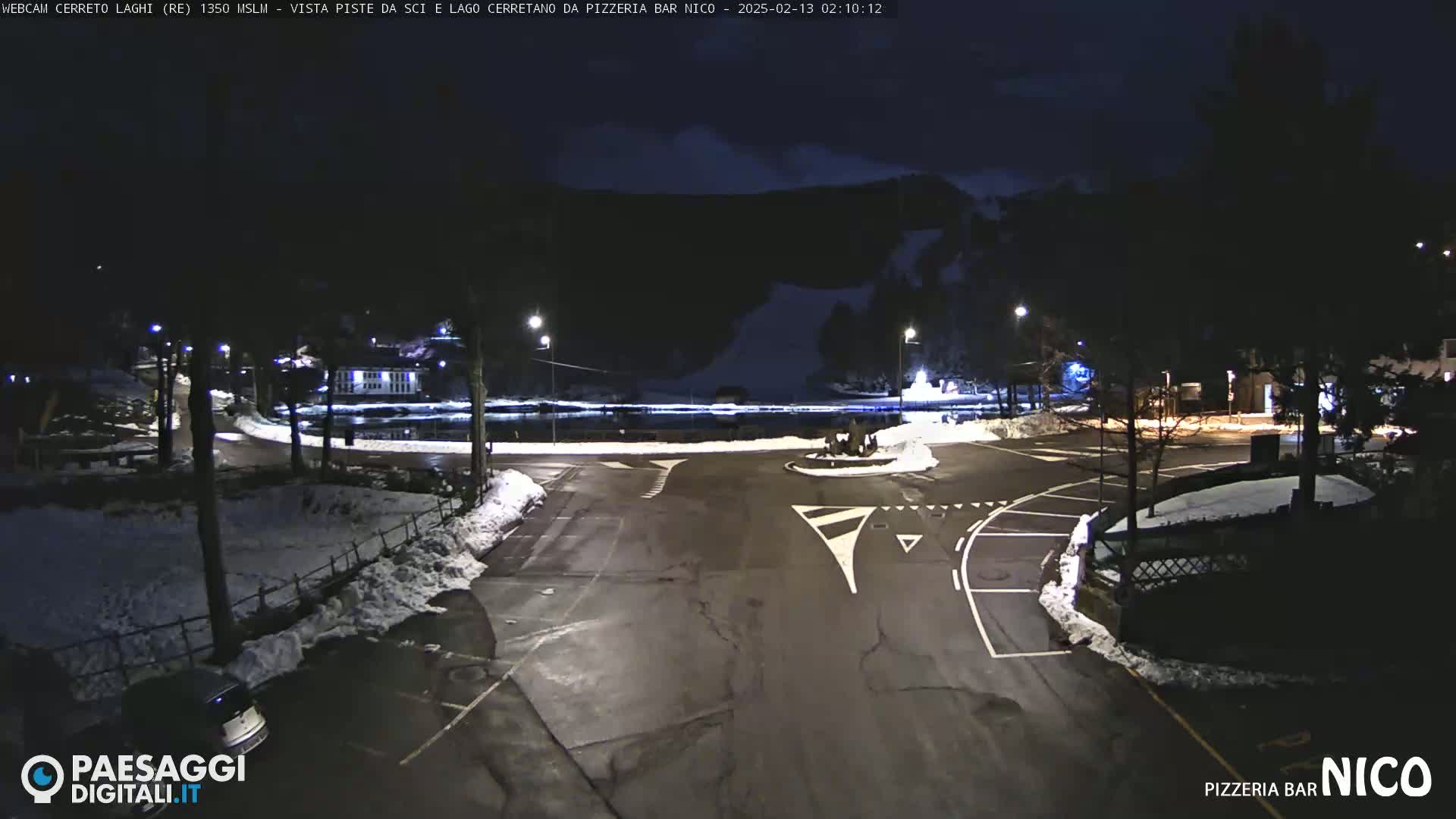 A nighttime view of a parking lot and roadway near a body of water, with snow on the ground and surrounding areas.
