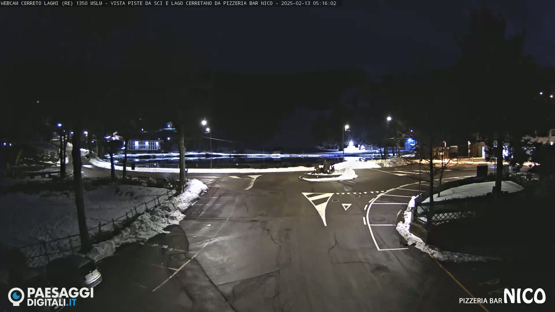 A nighttime view of a snow-dusted parking lot and roadway curving around a small body of water, illuminated by streetlights.