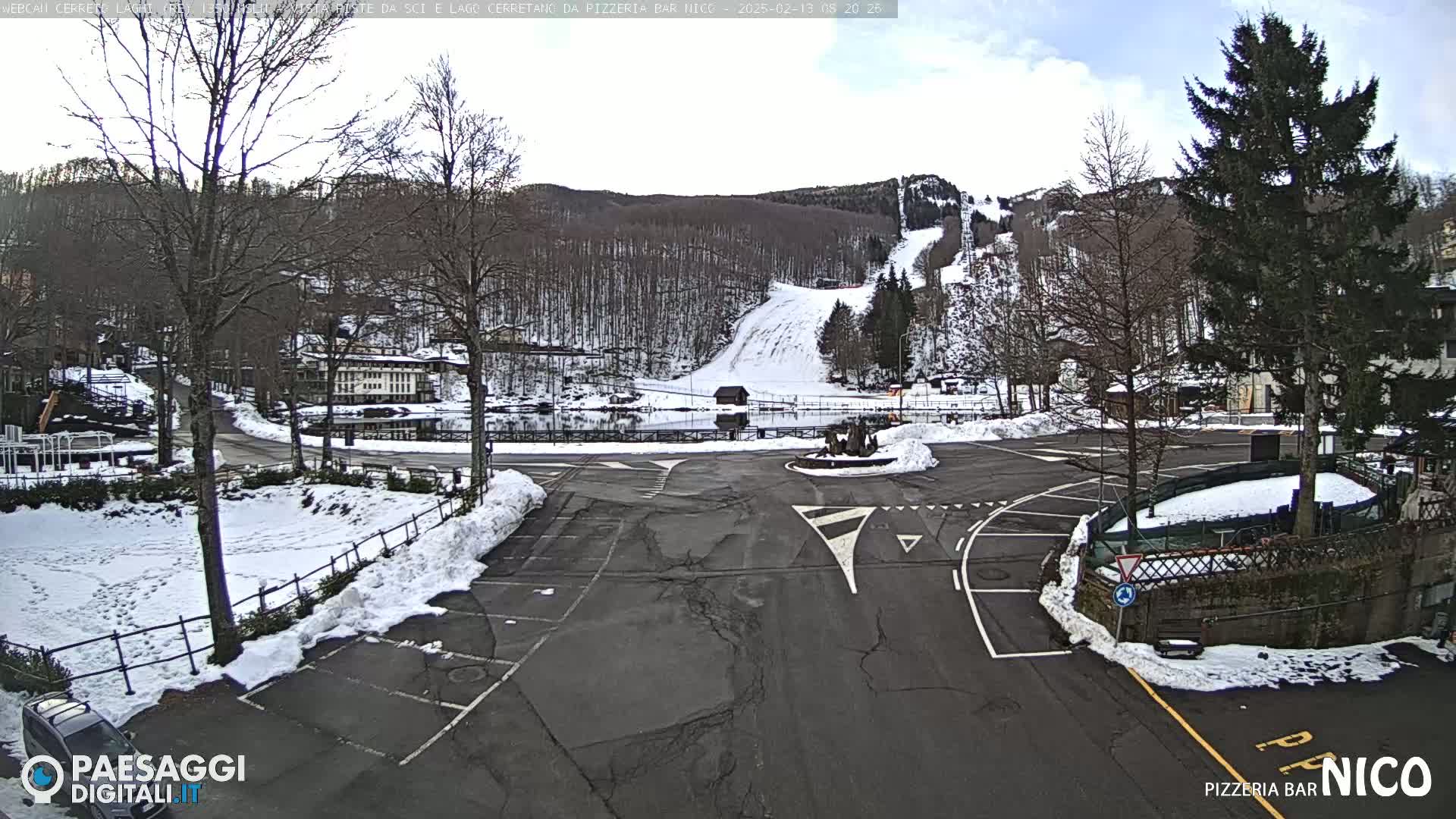 A snowy parking lot and a roundabout are in the foreground, with a ski slope and snow-covered trees visible in the background under a partly cloudy sky.