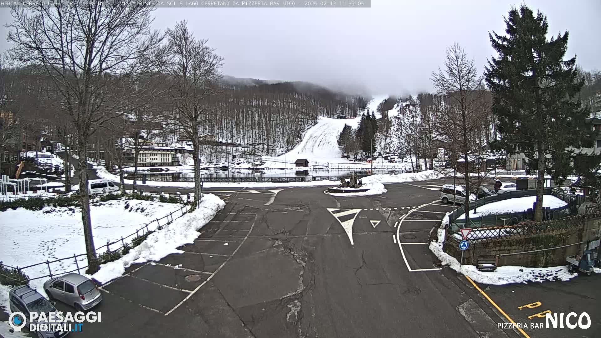A snow-covered parking lot and roadway are situated before a ski slope and a lake, with a misty, overcast sky overhead.