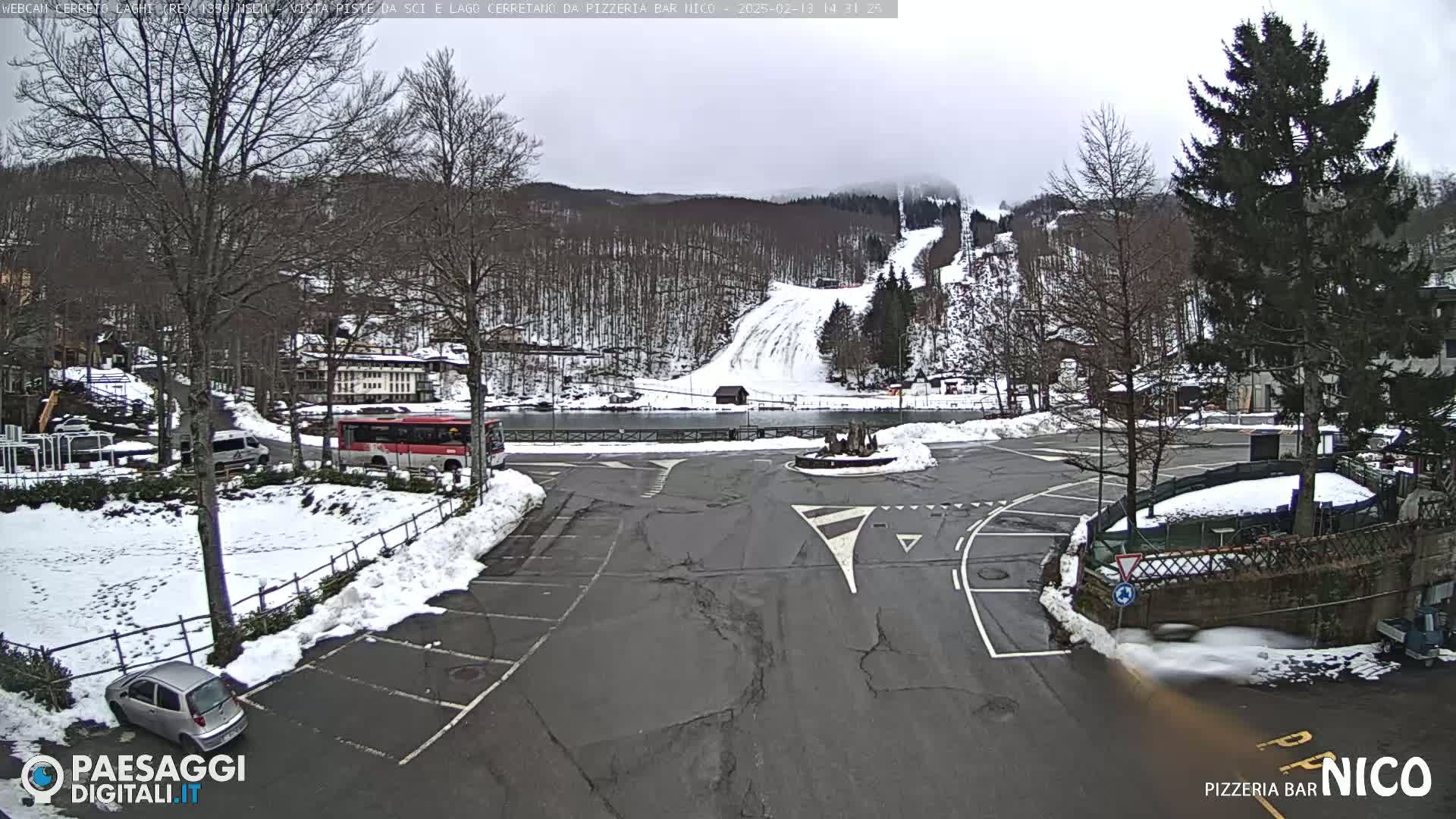 A snowy parking lot with a bus and cars, overlooks a ski slope on a cloudy day.