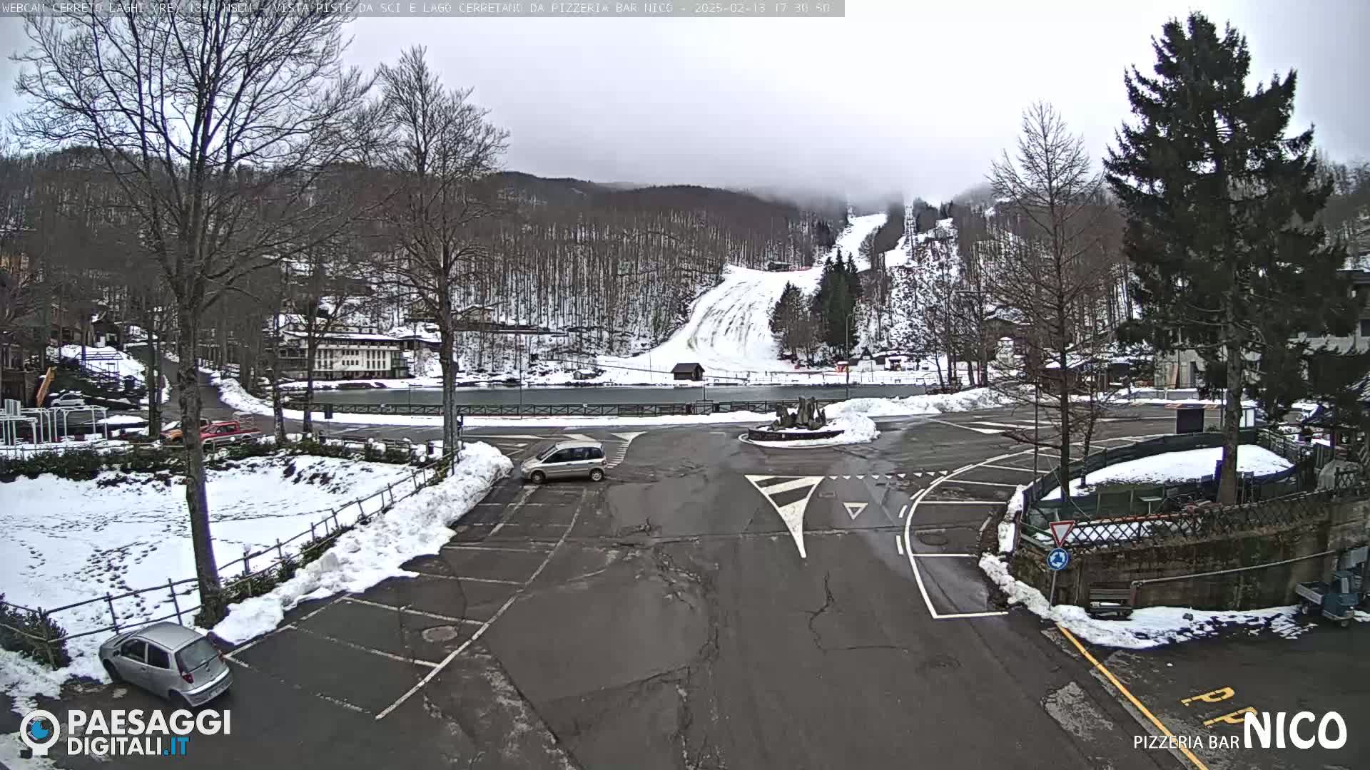 A mostly cloudy day shows a snow-covered parking lot and ski slope with several cars and buildings in the background.