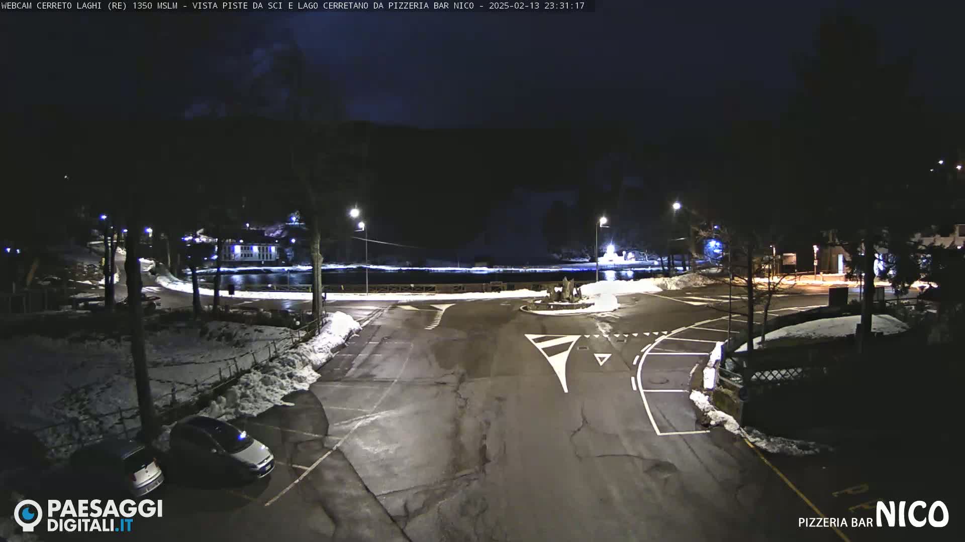 A nighttime view of a snow-dusted parking lot and roadway near a dark body of water, under a clear, dark sky.