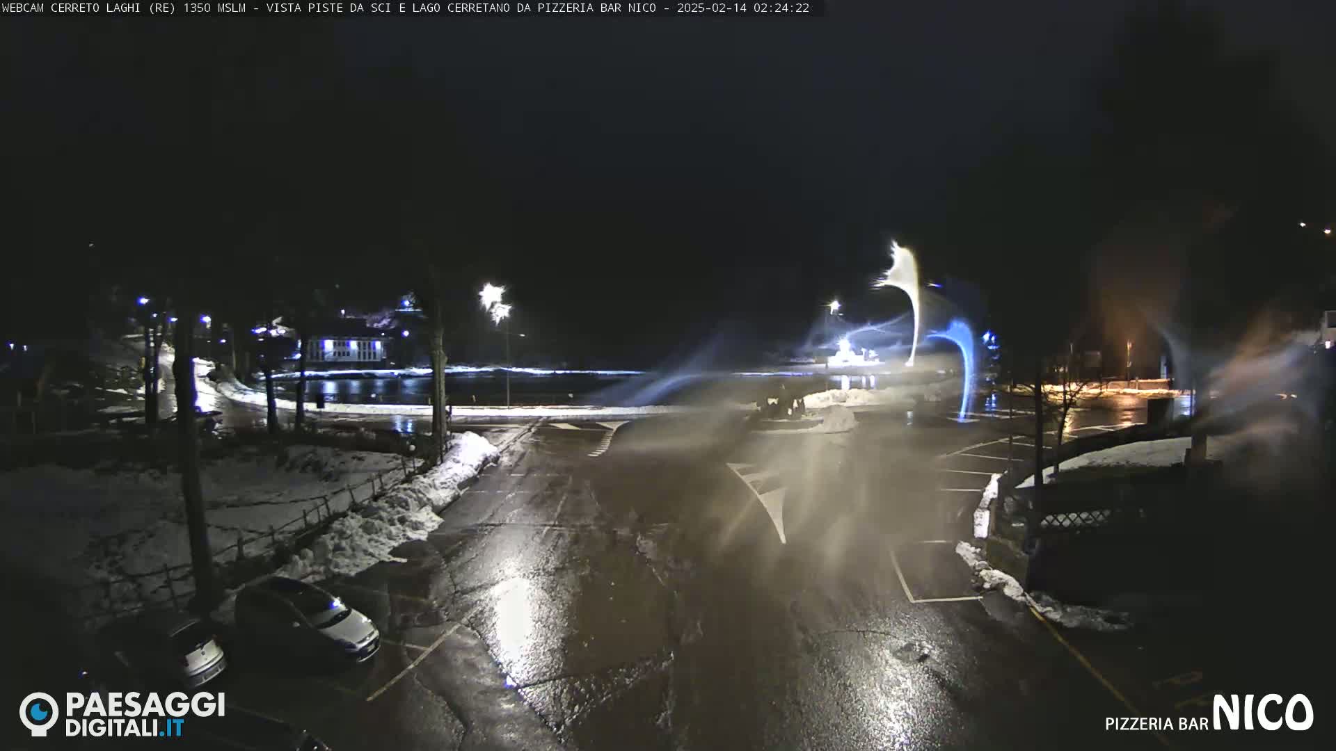 A nighttime scene of a snow-covered parking lot and road near a body of water, with snow blowing in the wind.