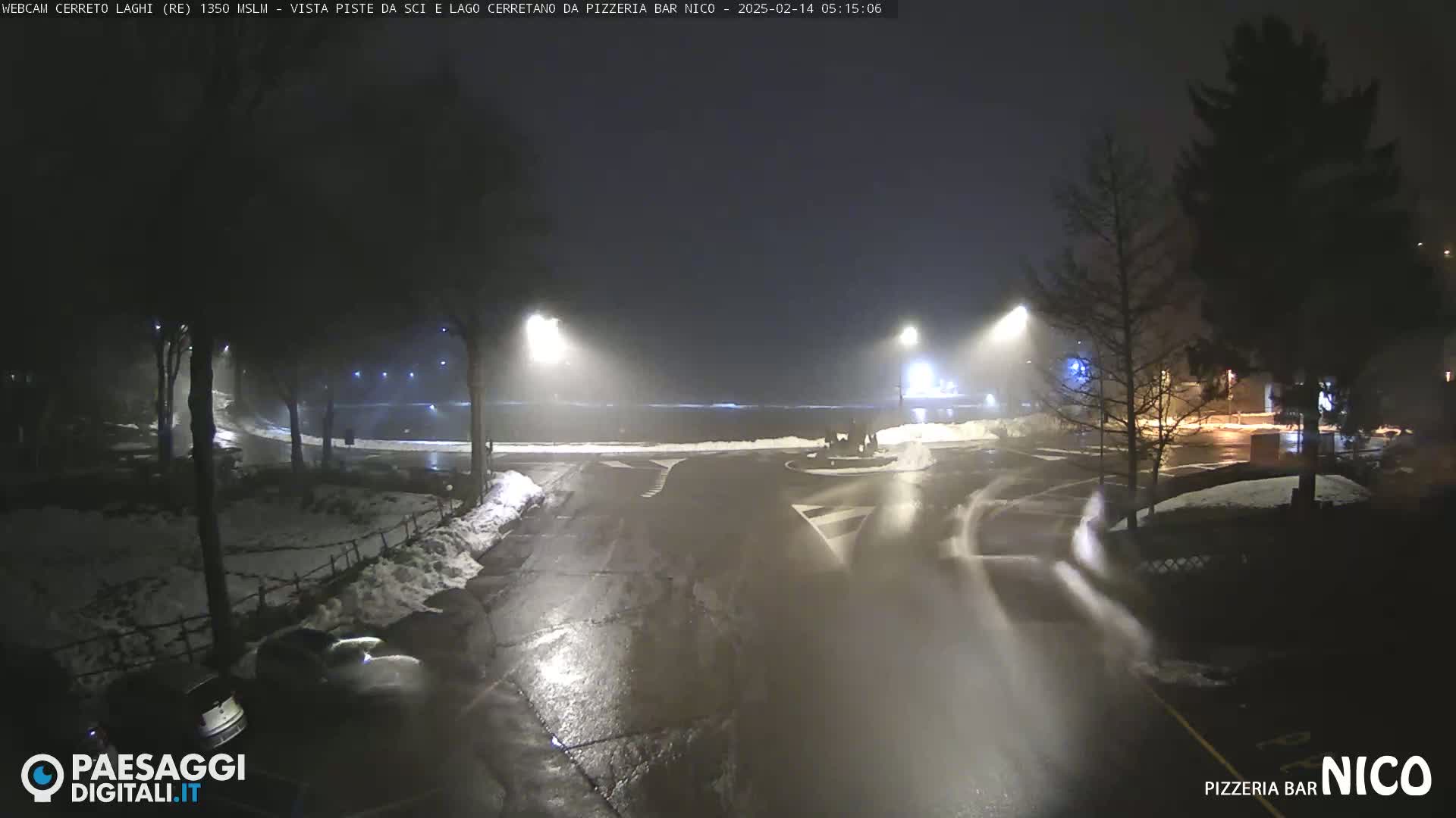 A nighttime, snowy, foggy scene shows a dark, wet road curving through a parking lot, with some cars visible and streetlights illuminating the area.