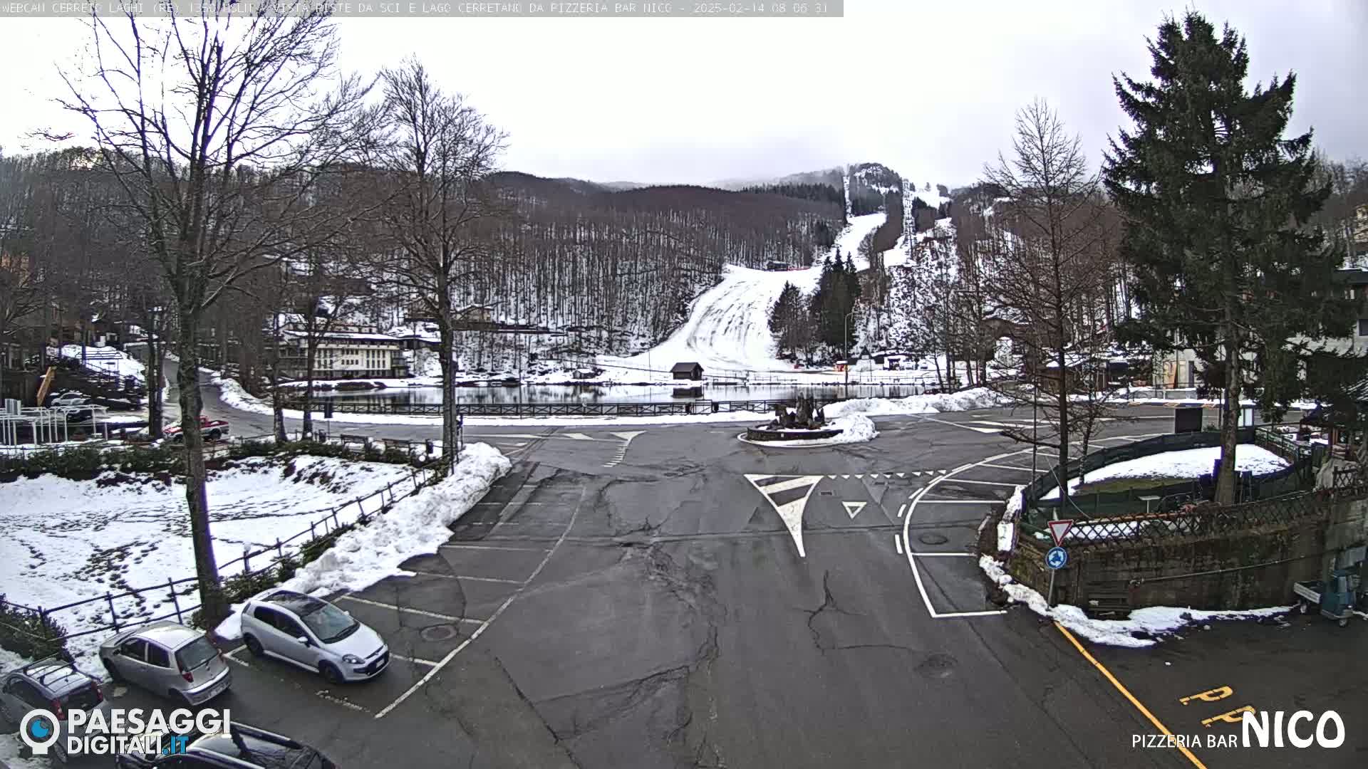 A parking lot with several cars, partially covered in snow, is situated in front of a small town nestled at the base of a snow-covered ski slope and wooded mountain under an overcast sky.