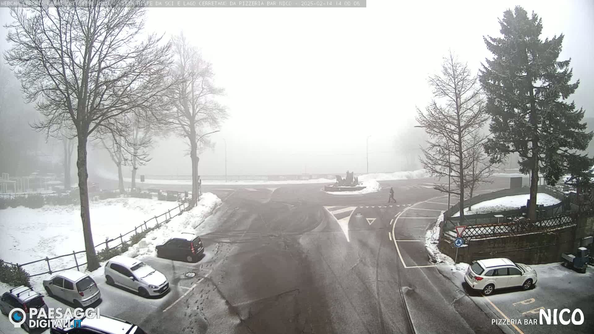 A snow-covered parking lot and road intersection are shrouded in fog, with several cars parked and a pedestrian walking.