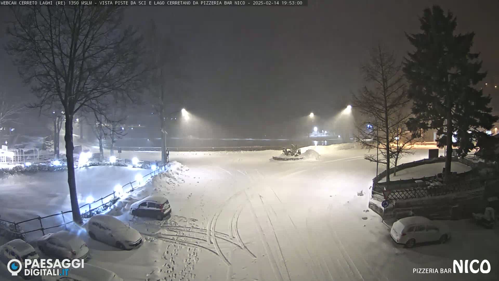 A snow-covered parking lot at night, illuminated by streetlights, with several cars parked and tire tracks visible in the snow.