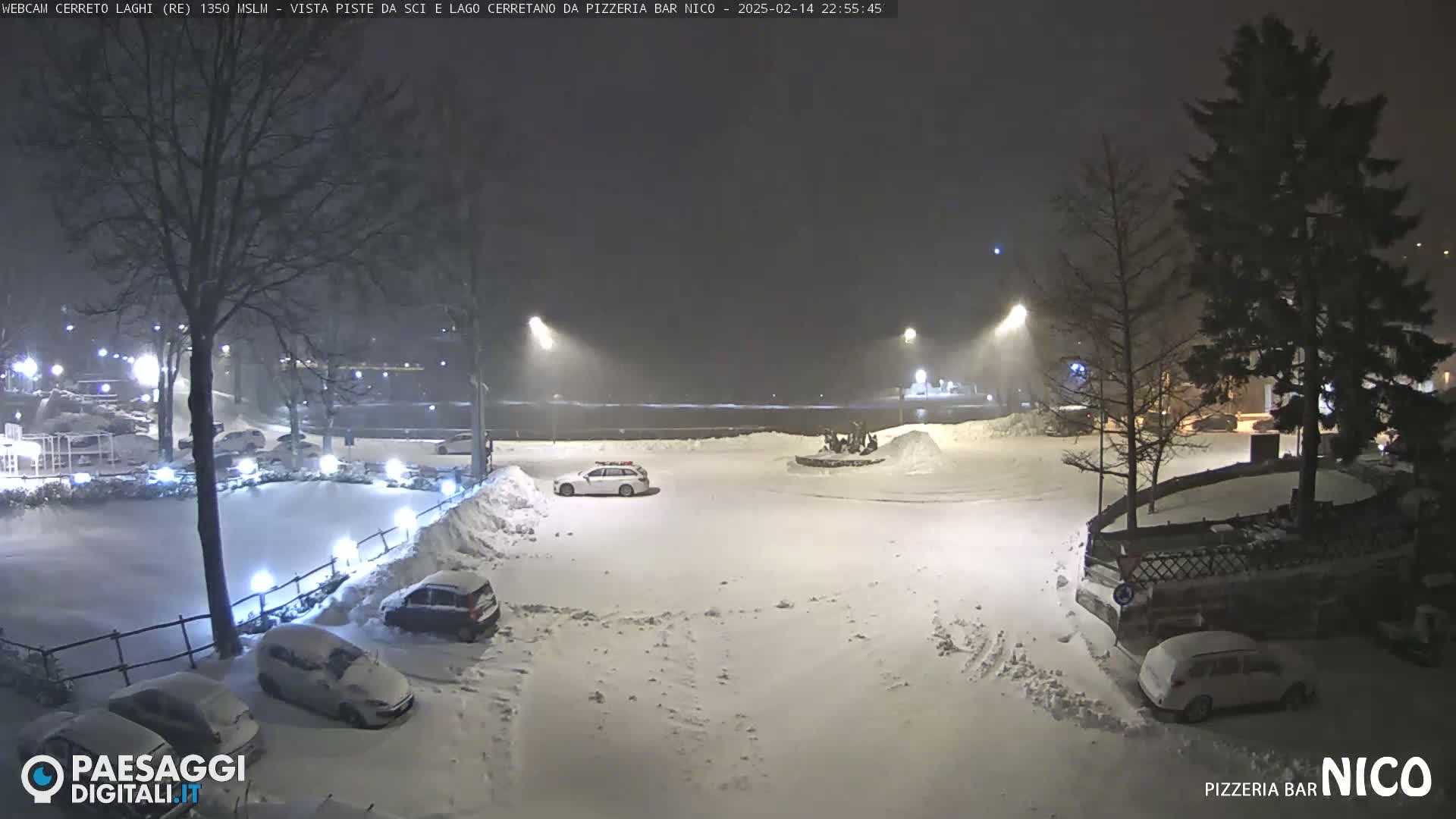 A snow-covered parking lot at night, illuminated by streetlights, with several cars parked and a few people gathered near a circular structure in the background under a heavy snowfall.