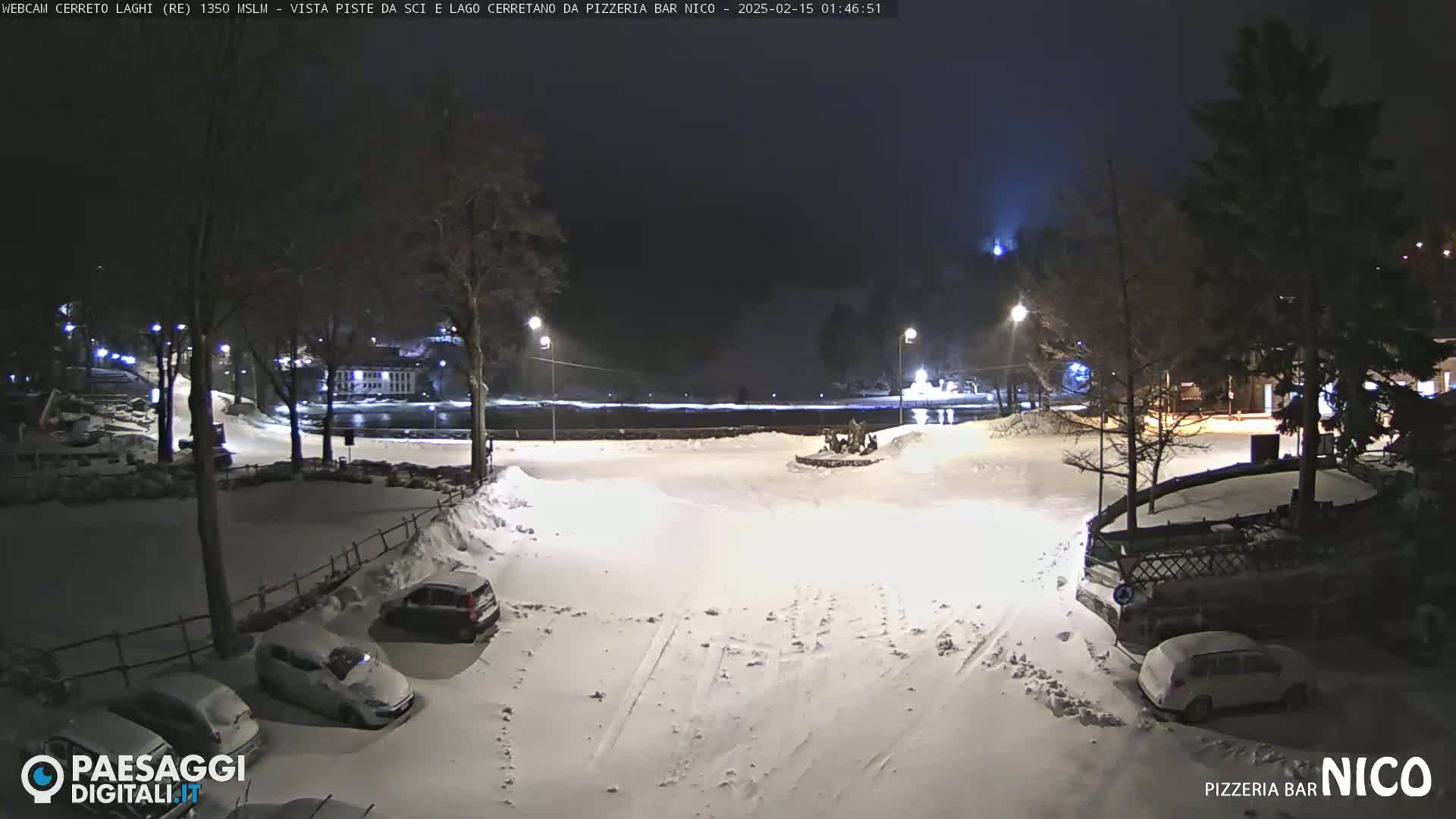A snow-covered nighttime scene shows a parking lot with several cars, a body of water in the background, and trees lining the perimeter under a dark sky.