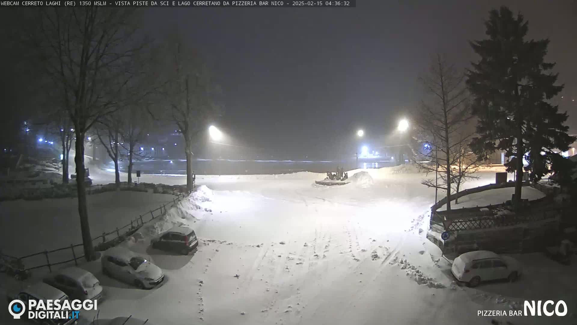 A snow-covered parking lot at night, illuminated by streetlights, with several cars parked and a few trees in the background.