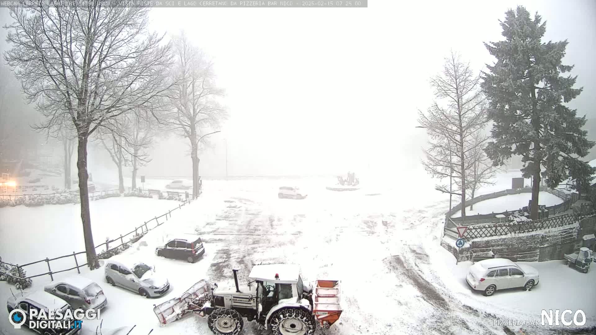 A snow-covered parking lot and road with several cars and a snowplow are visible in heavy snowfall.
