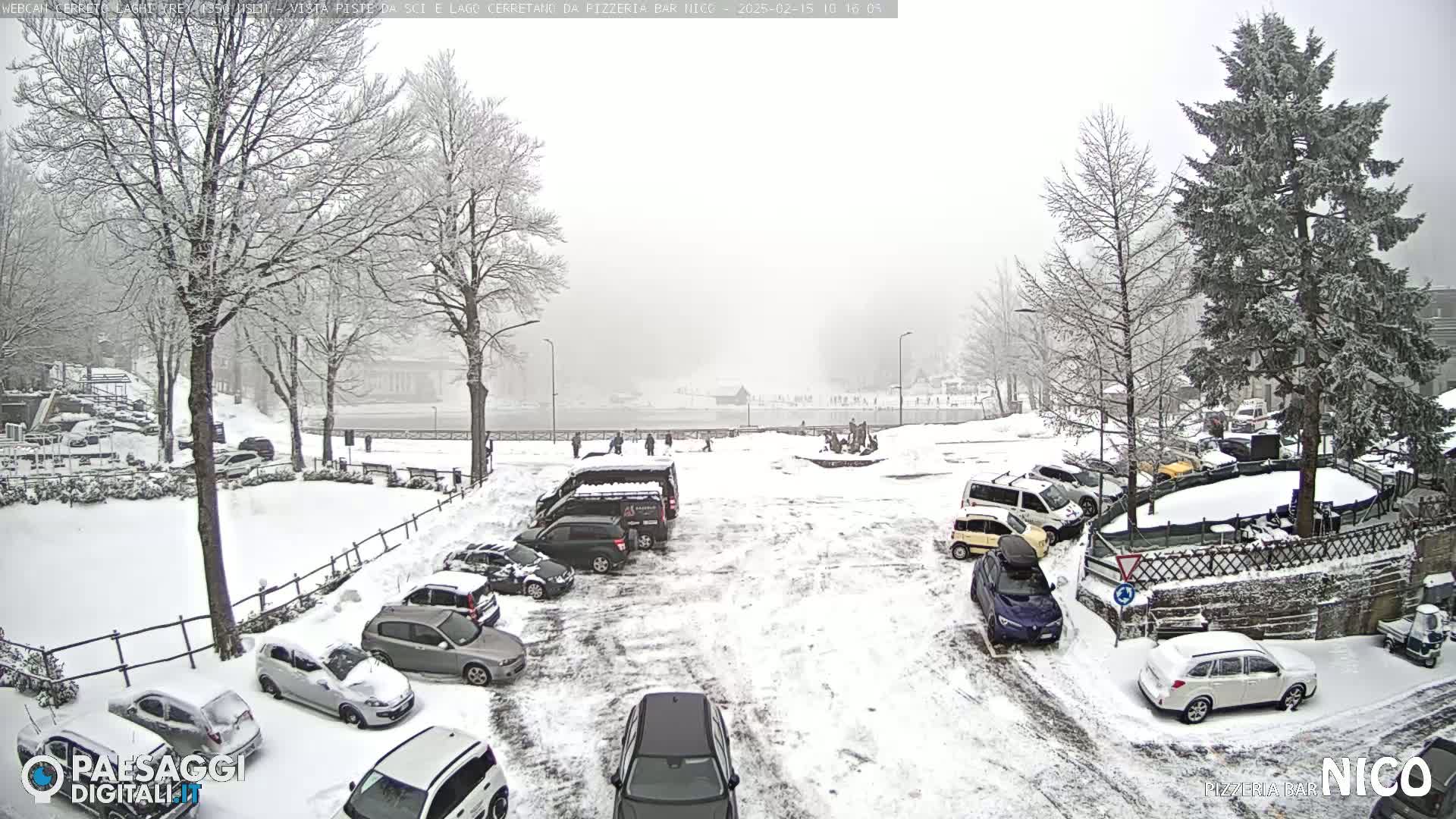 A snow-covered parking lot with cars and snow-laden trees surrounds a partially visible body of water under a heavily overcast sky.